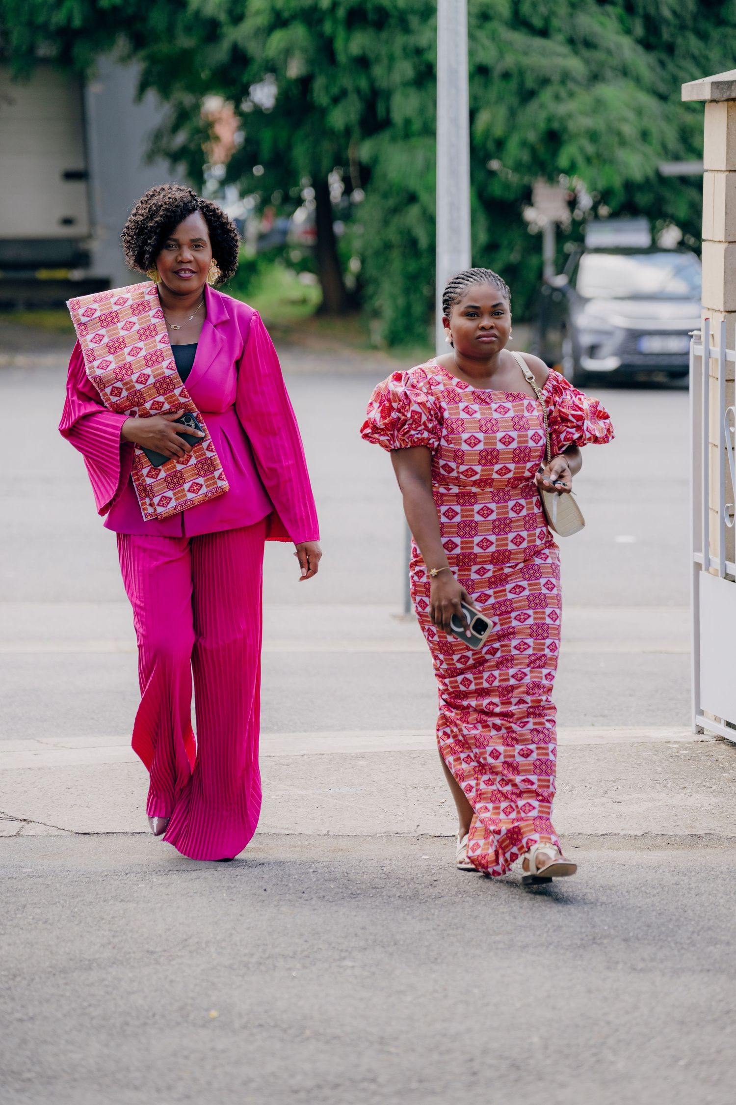 Group wearing bright pink and patterned red dresses walking in a parking area.