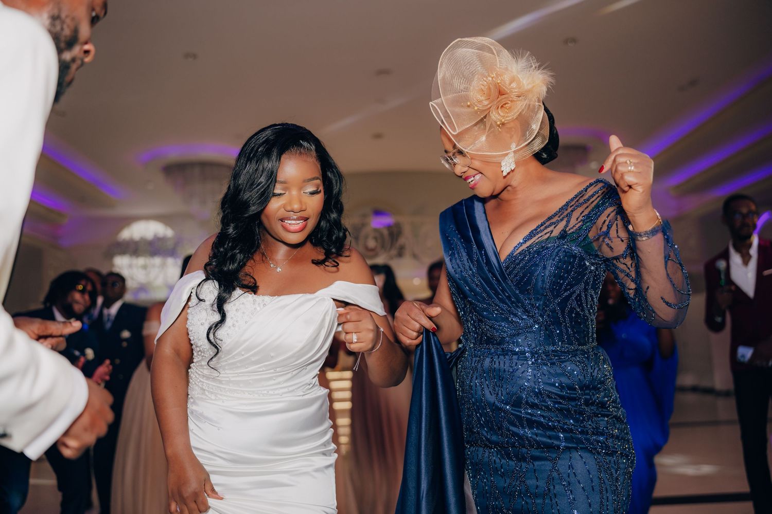 Two women in elegant dresses, one white and one navy blue, dance together at a wedding reception.