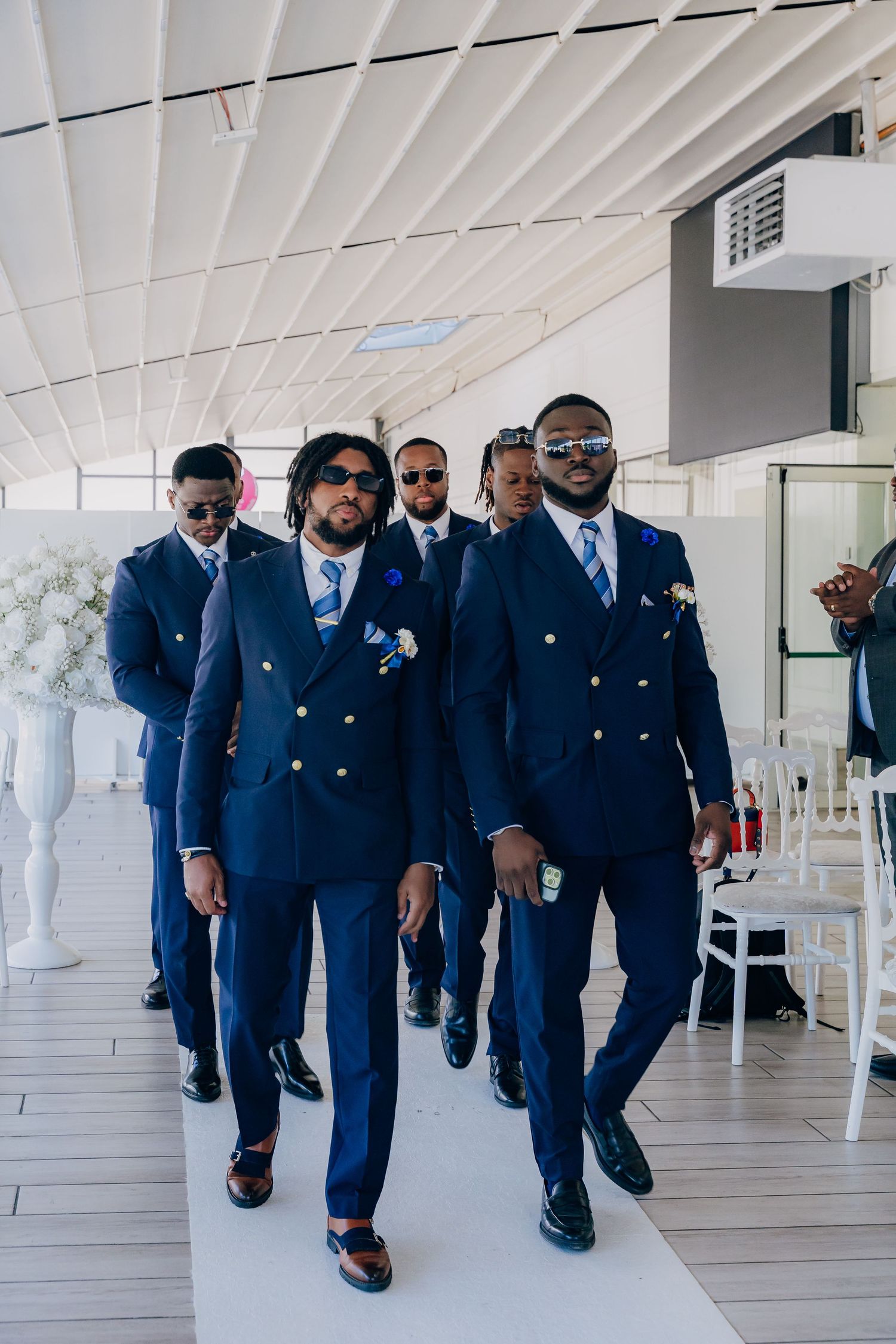 Groomsmen in matching navy suits walk down a white-decorated hallway at a formal event.