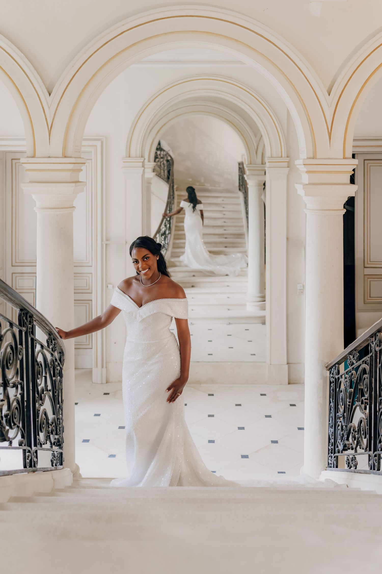 Elegant bride in off-shoulder white gown poses in grand hallway with arched columns and ornate iron railings.