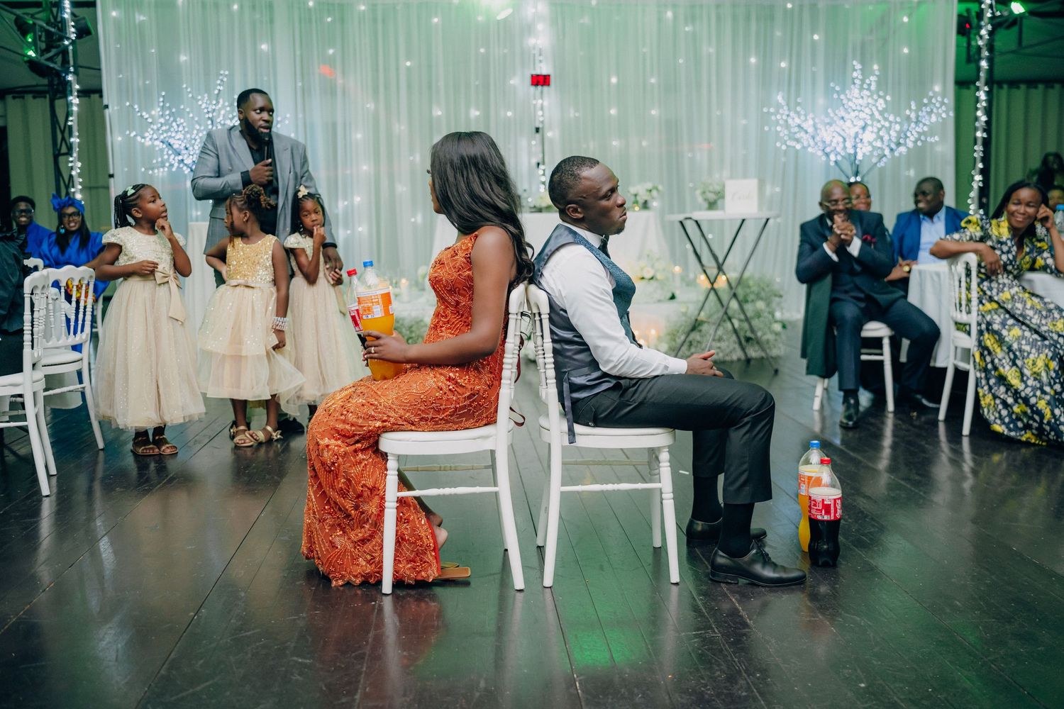 Wedding reception scene with guests seated at tables under twinkling green lights and white decor.