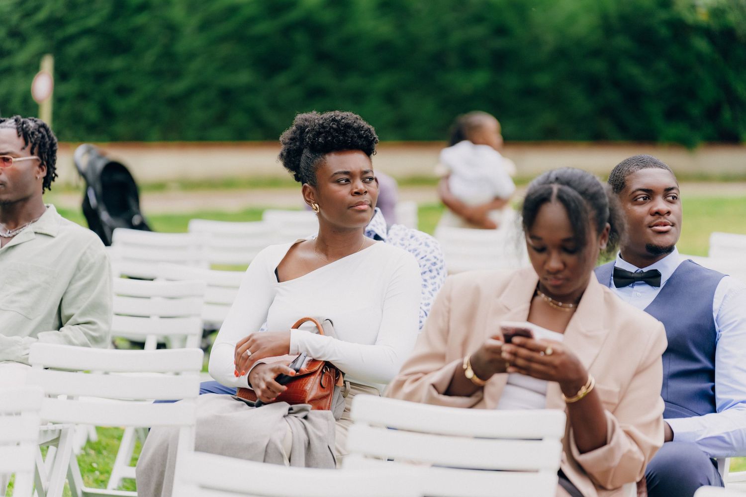 Wedding guests seated in white chairs outdoors check their phones during a ceremony.
