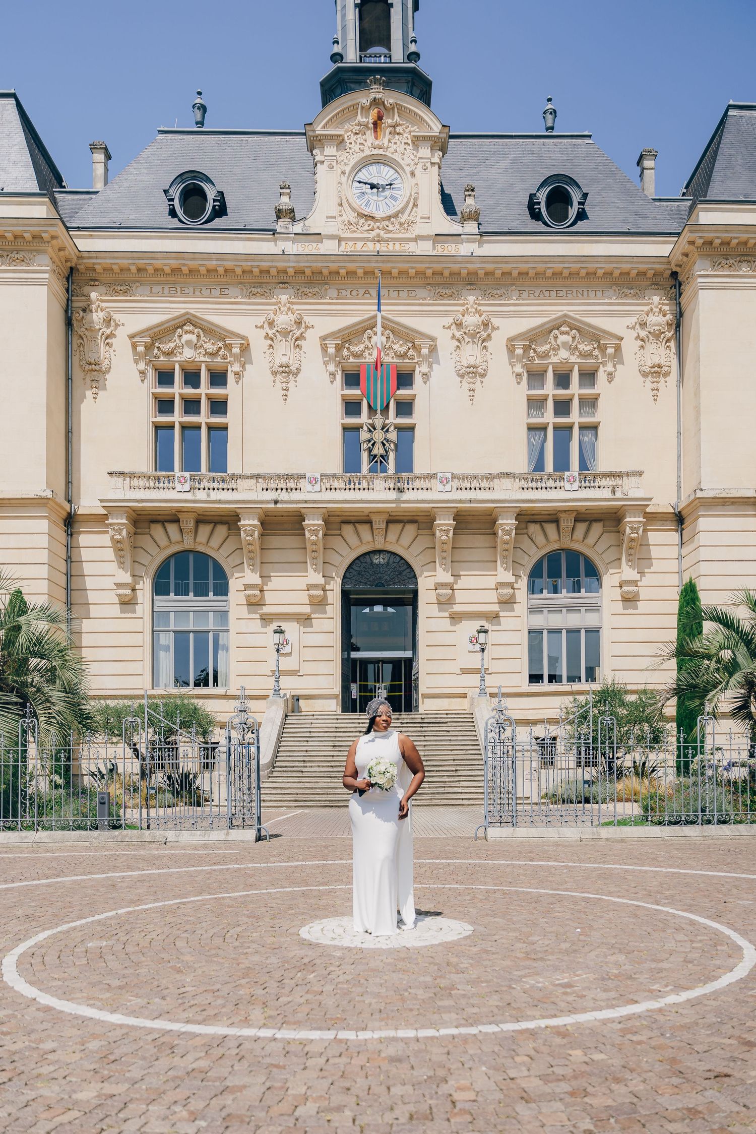 Elegant French chateau building with ornate architecture serves as backdrop for figure in white dress standing in circular courtyard.