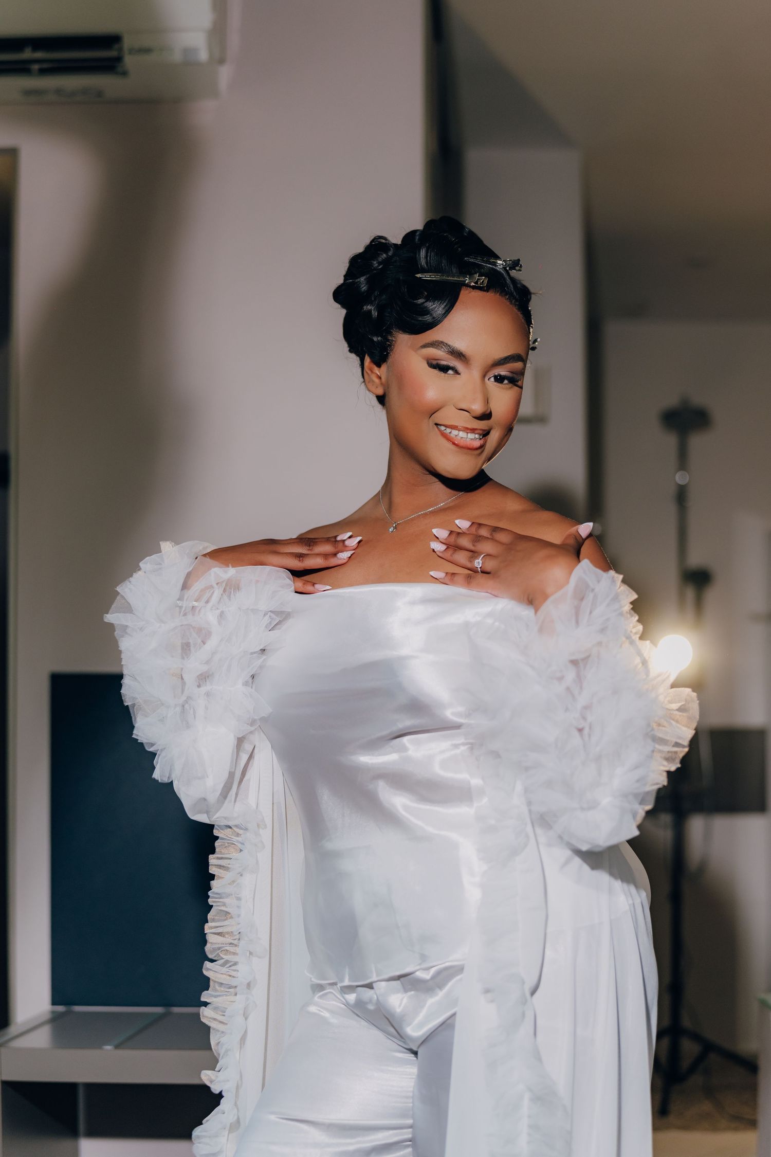 A person wearing an elegant white wedding dress with ruffled off-shoulder sleeves smiles while showing off their ring.