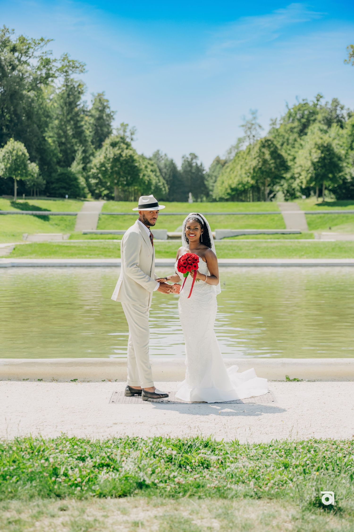 Two elegantly dressed people stand together by a pond in a scenic park on a bright summer day.