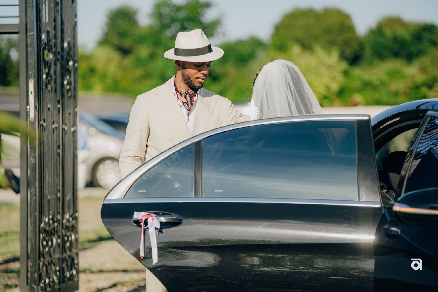 A well-dressed couple in light-colored attire stands near a black car on a sunny summer day.