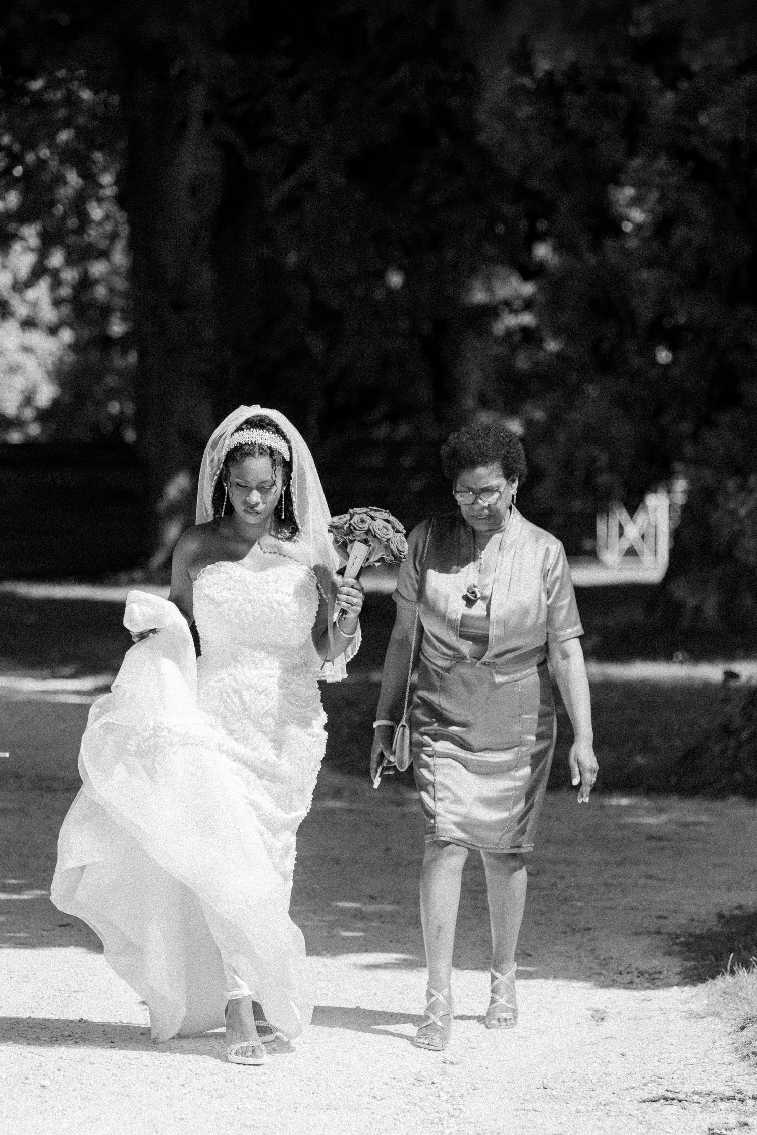 A bride in a white wedding gown walks alongside a woman in a grey dress on a garden path, captured in black and white.