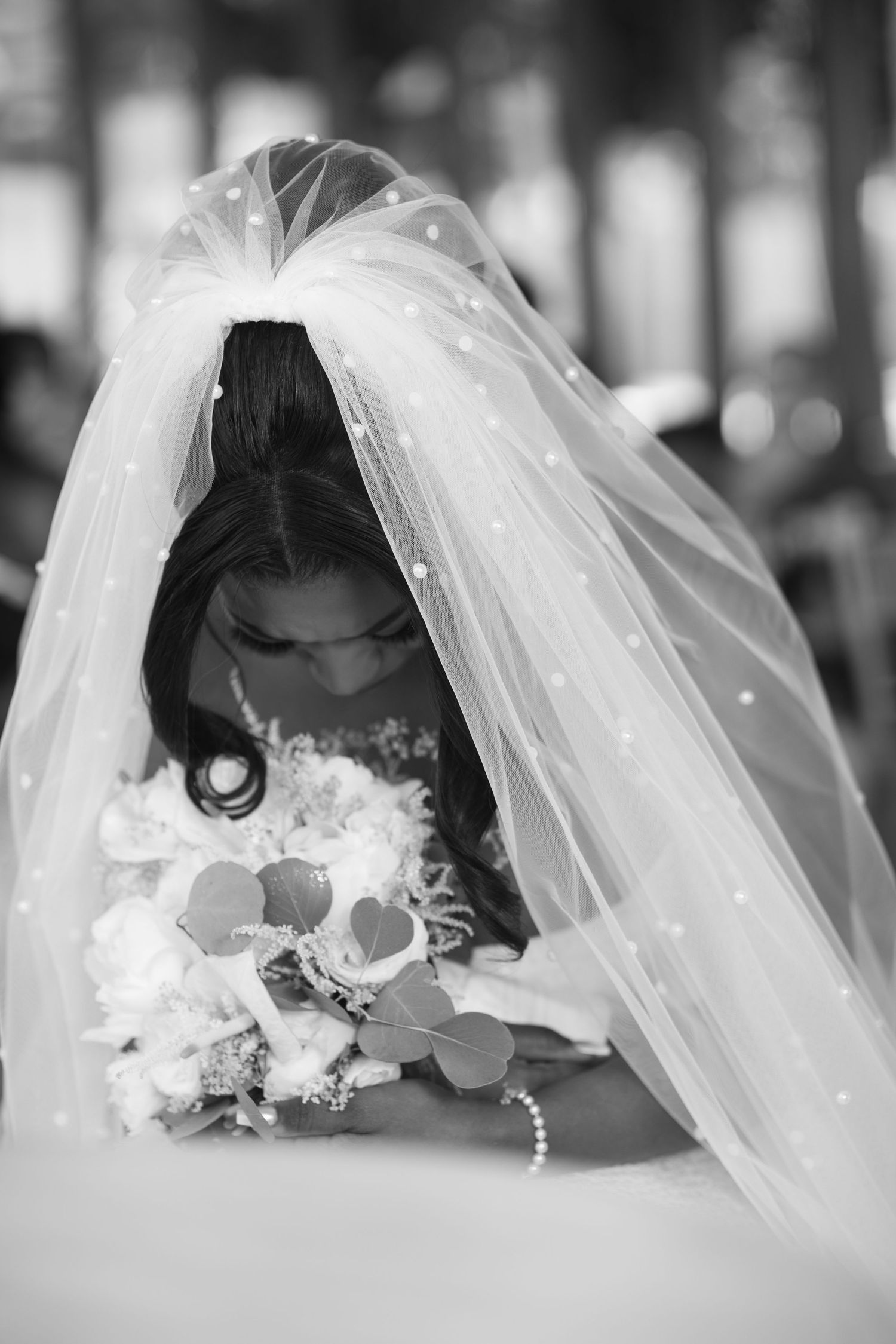 Black and white photo of a bride bowing her head under a flowing veil with white flowers.