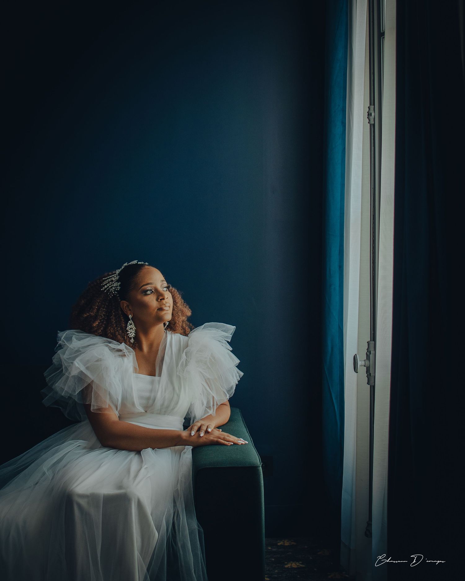 A bride wearing an elegant white tulle dress sits contemplatively by a window against a deep blue wall in dramatic lighting.