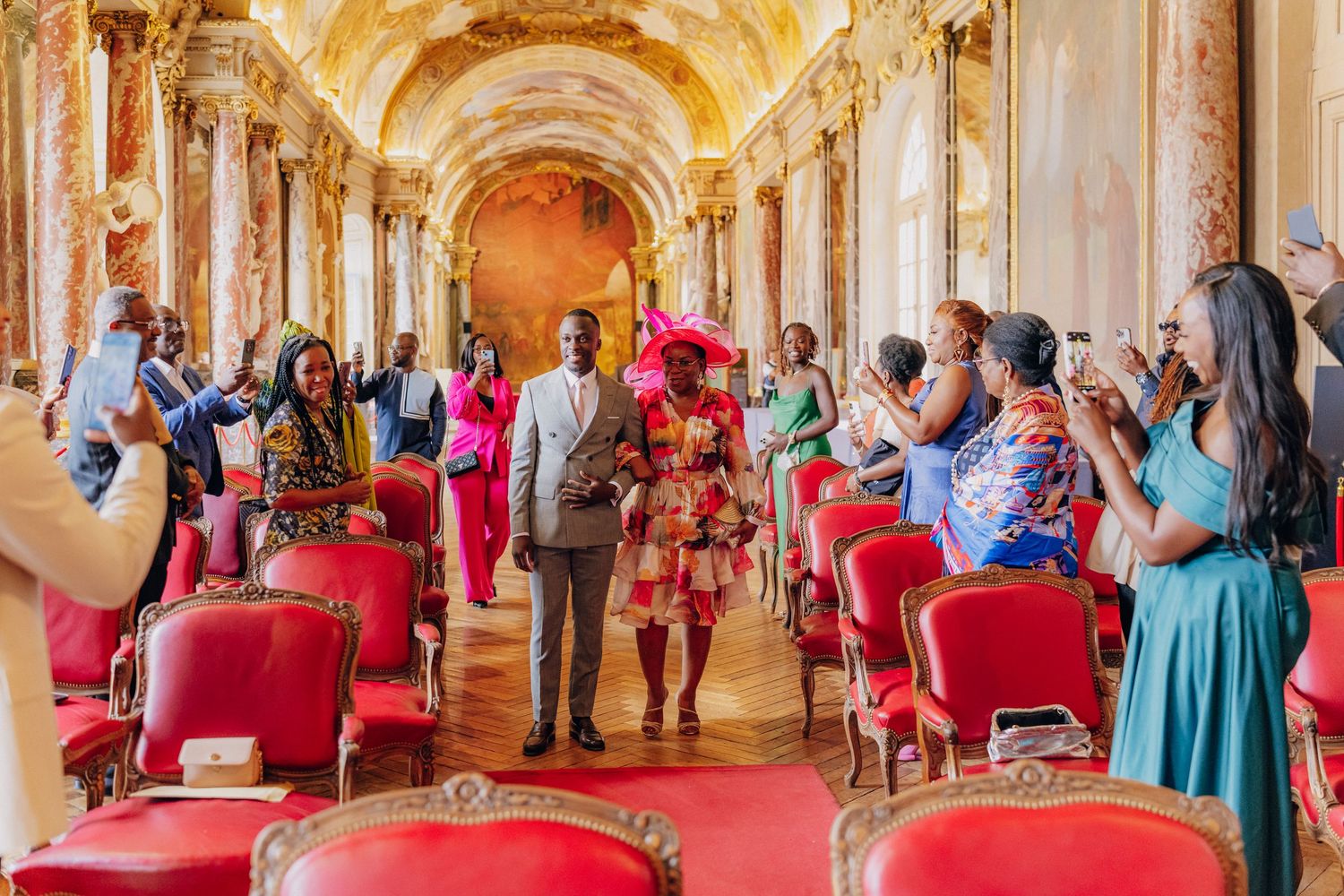 Ornate baroque palace interior hallway with red chairs and tourists viewing gilded architectural details and frescoes.