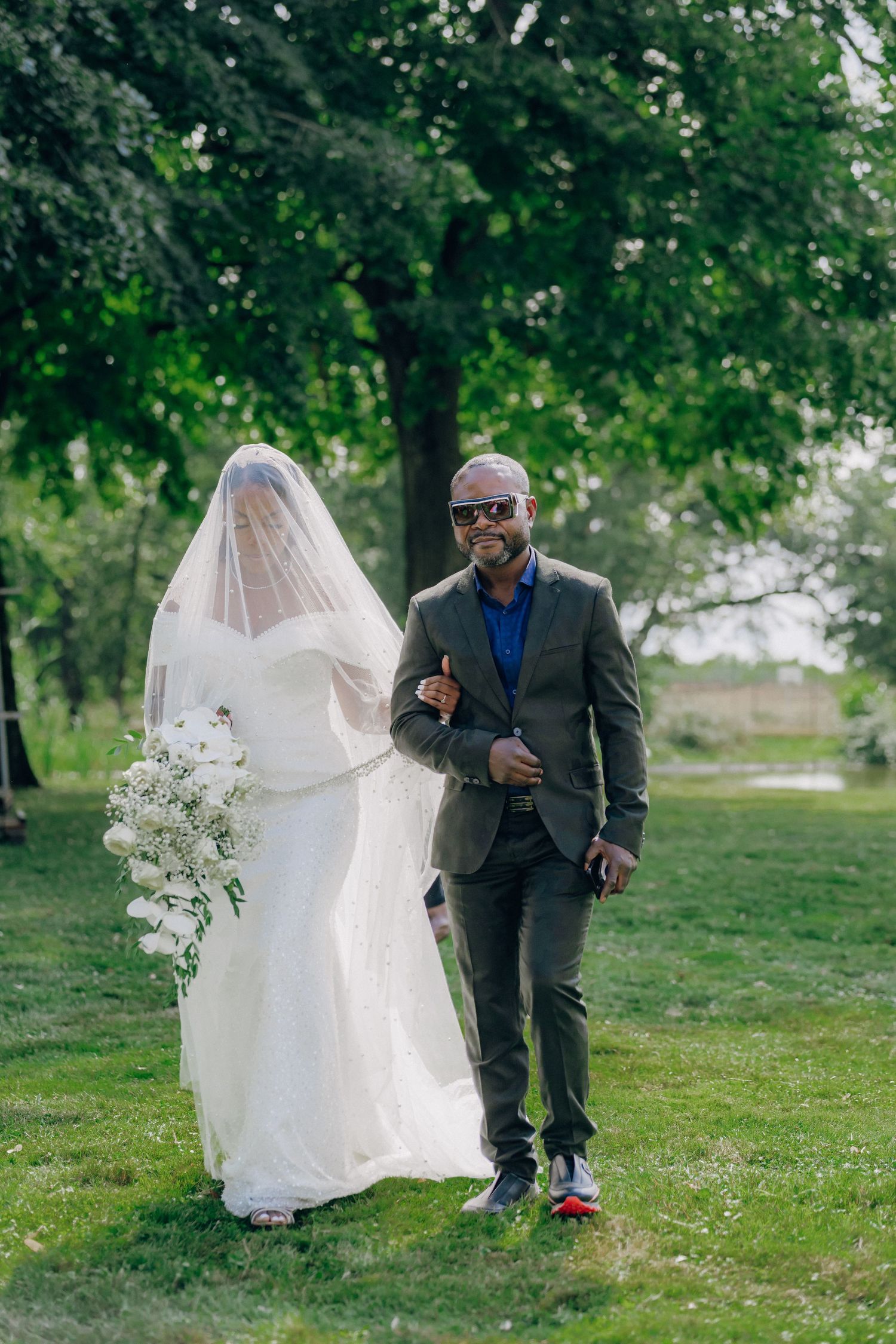 A bride in a flowing white dress walks across a grassy lawn with a bouquet of white flowers.