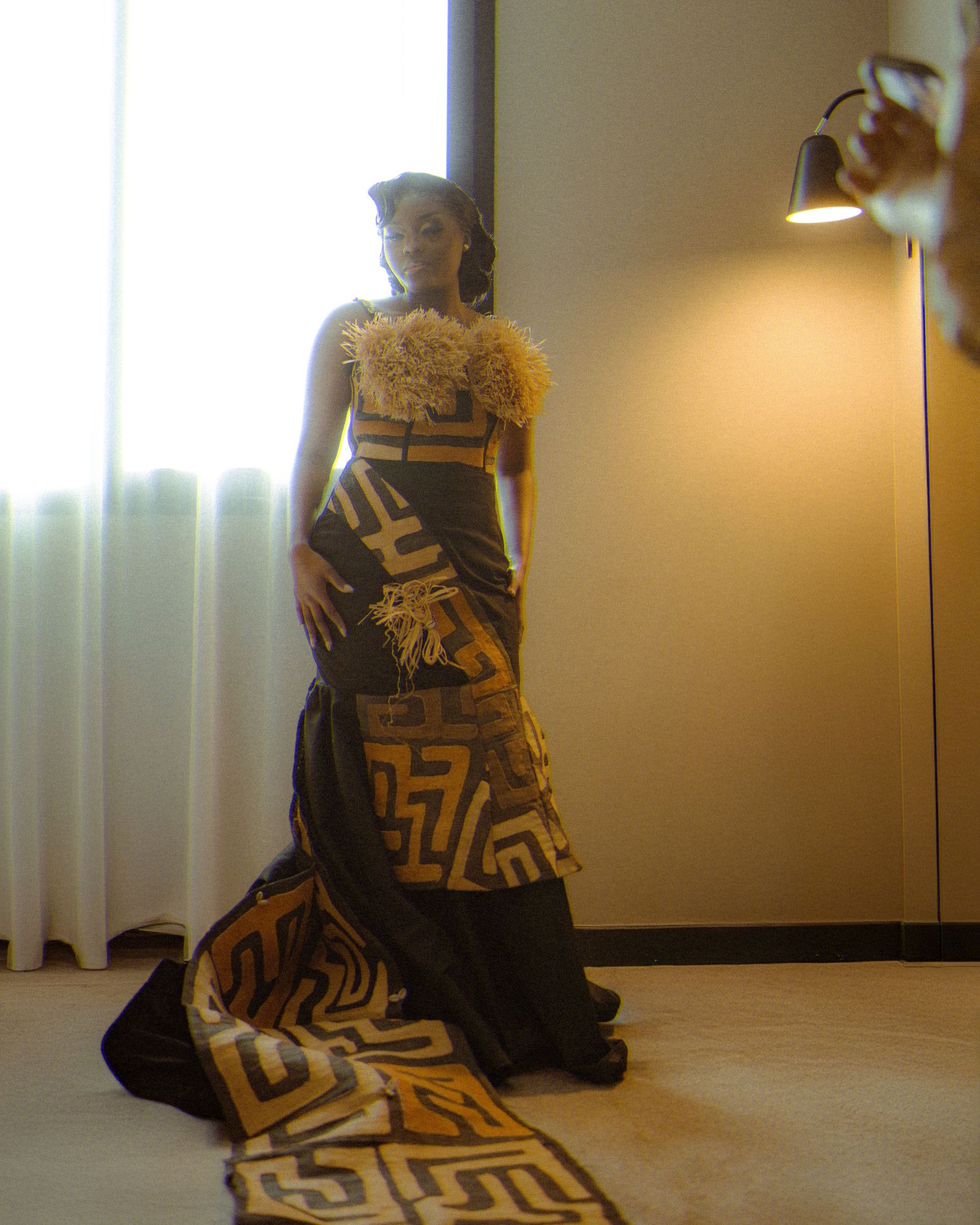 Person wearing an elegant black and gold evening gown with geometric patterns stands in dramatic lighting by a window.