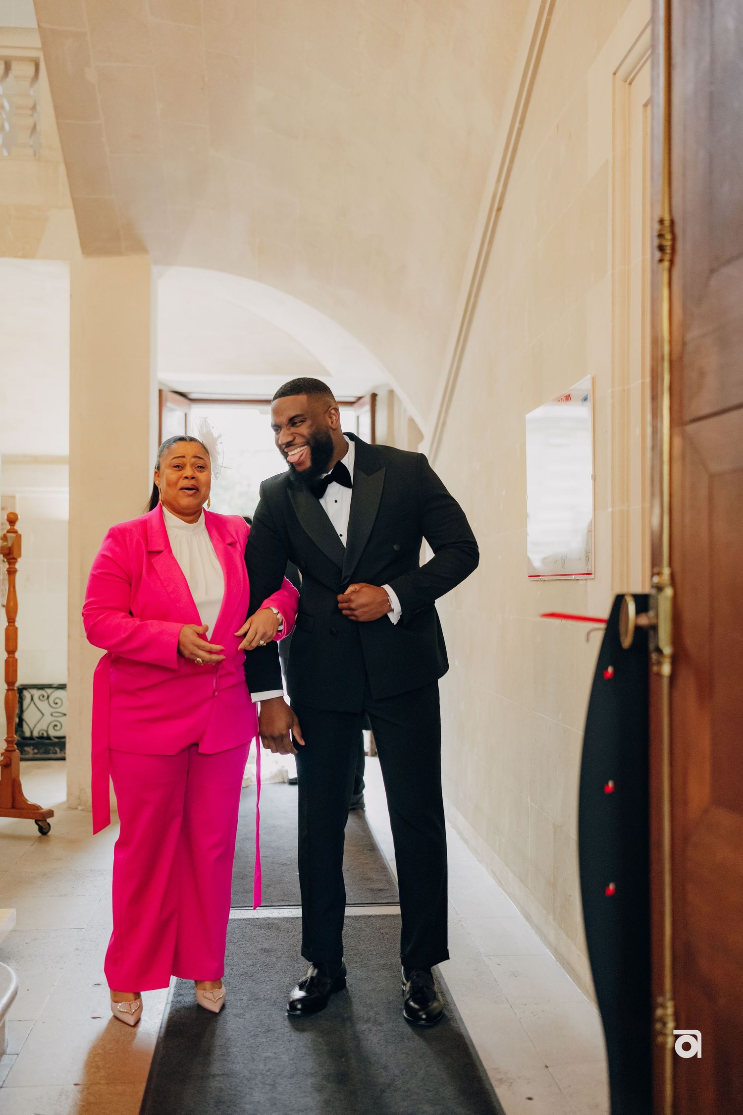 Two people walk down an elegant hallway, with one wearing a bright pink suit and the other in a classic black tuxedo.