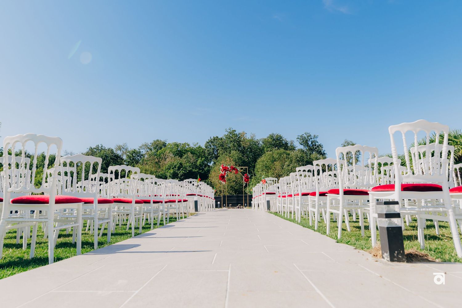 Rows of white chairs line an outdoor wedding ceremony aisle with red ribbons under a bright blue sky.