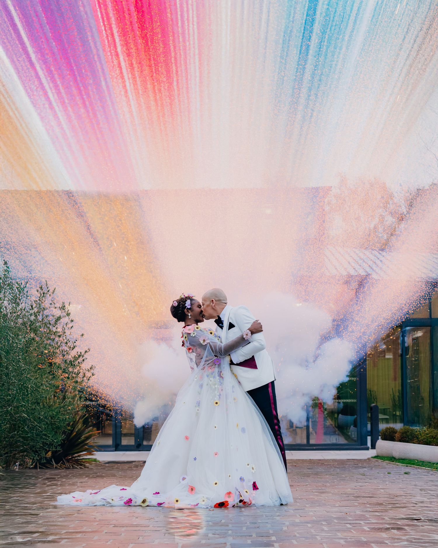 A couple in wedding attire dances surrounded by colorful smoke bombs creating a dreamy pink and orange atmosphere.