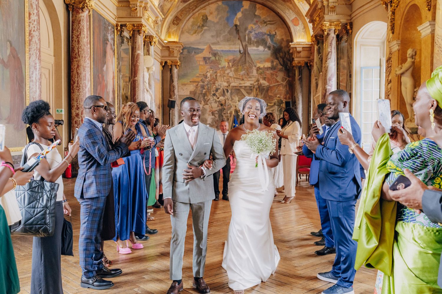 Wedding guests gathered in a grand palace gallery with decorative murals and arched ceilings.