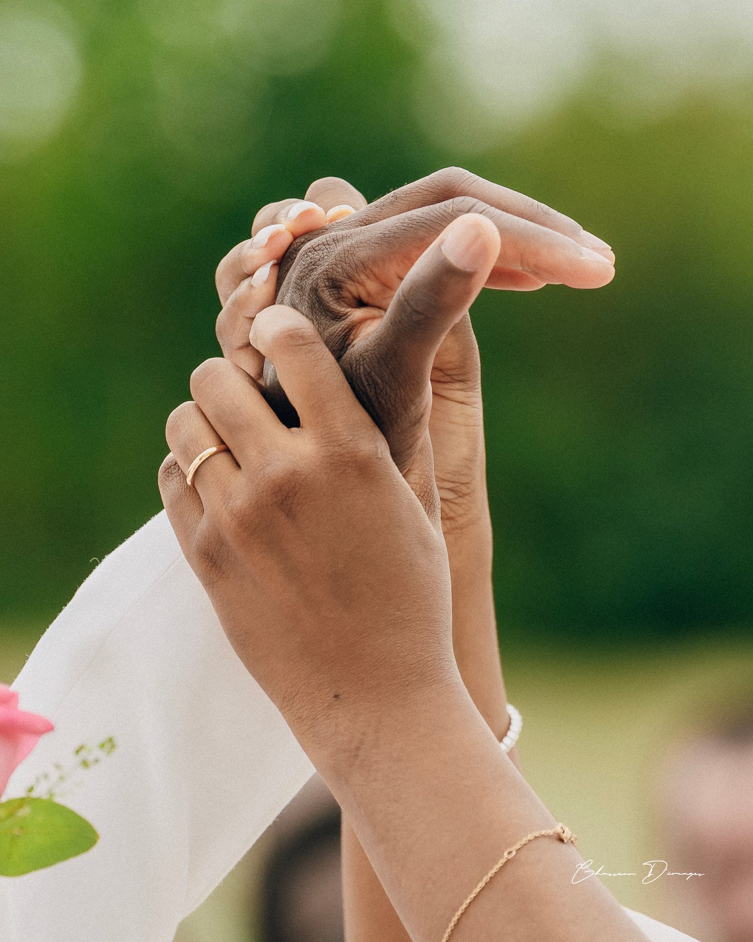 Intimate close-up of intertwined hands showing wedding rings against blurred green foliage.