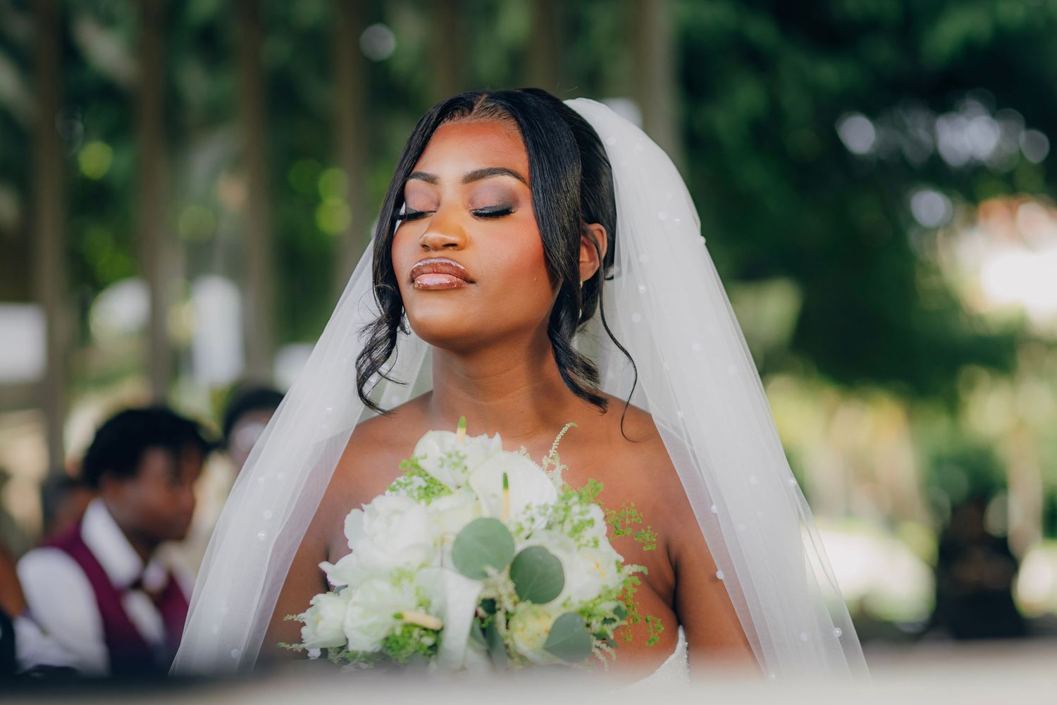 A bride in a white gown holds a bouquet of white flowers while wearing a flowing veil outdoors.