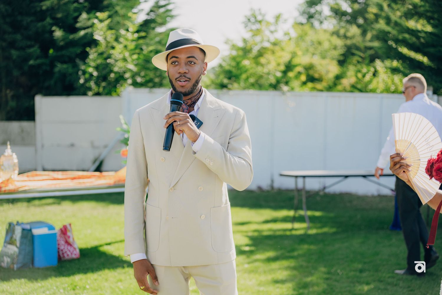 A person in a cream-colored suit and cap speaks into a microphone at an outdoor event on a sunny day.