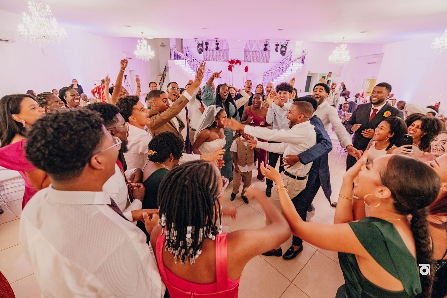 A lively wedding reception dance party with guests raising their arms on a crowded dance floor under pink lighting.