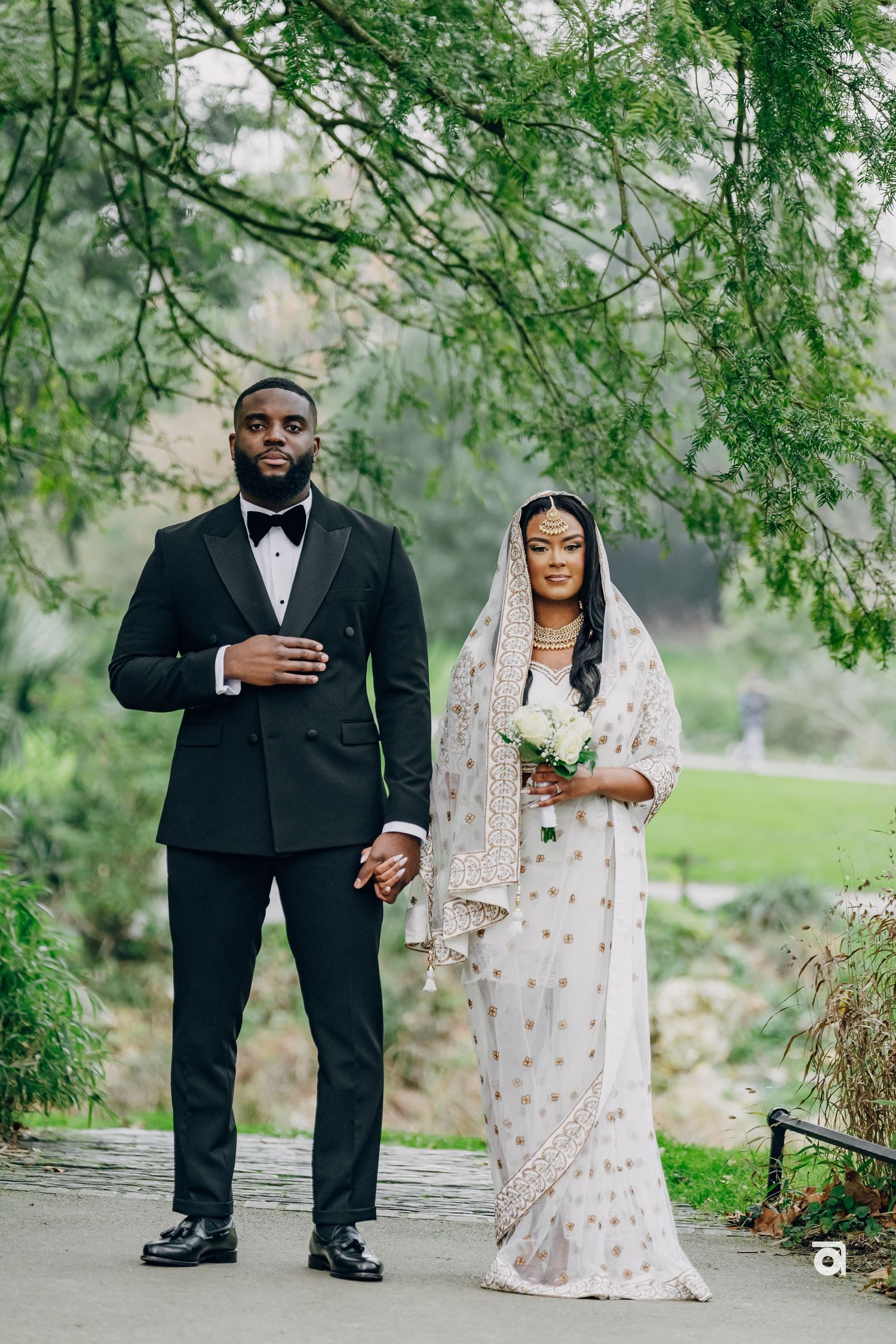 A couple in formal wedding attire stands beneath hanging tree branches on a garden pathway.