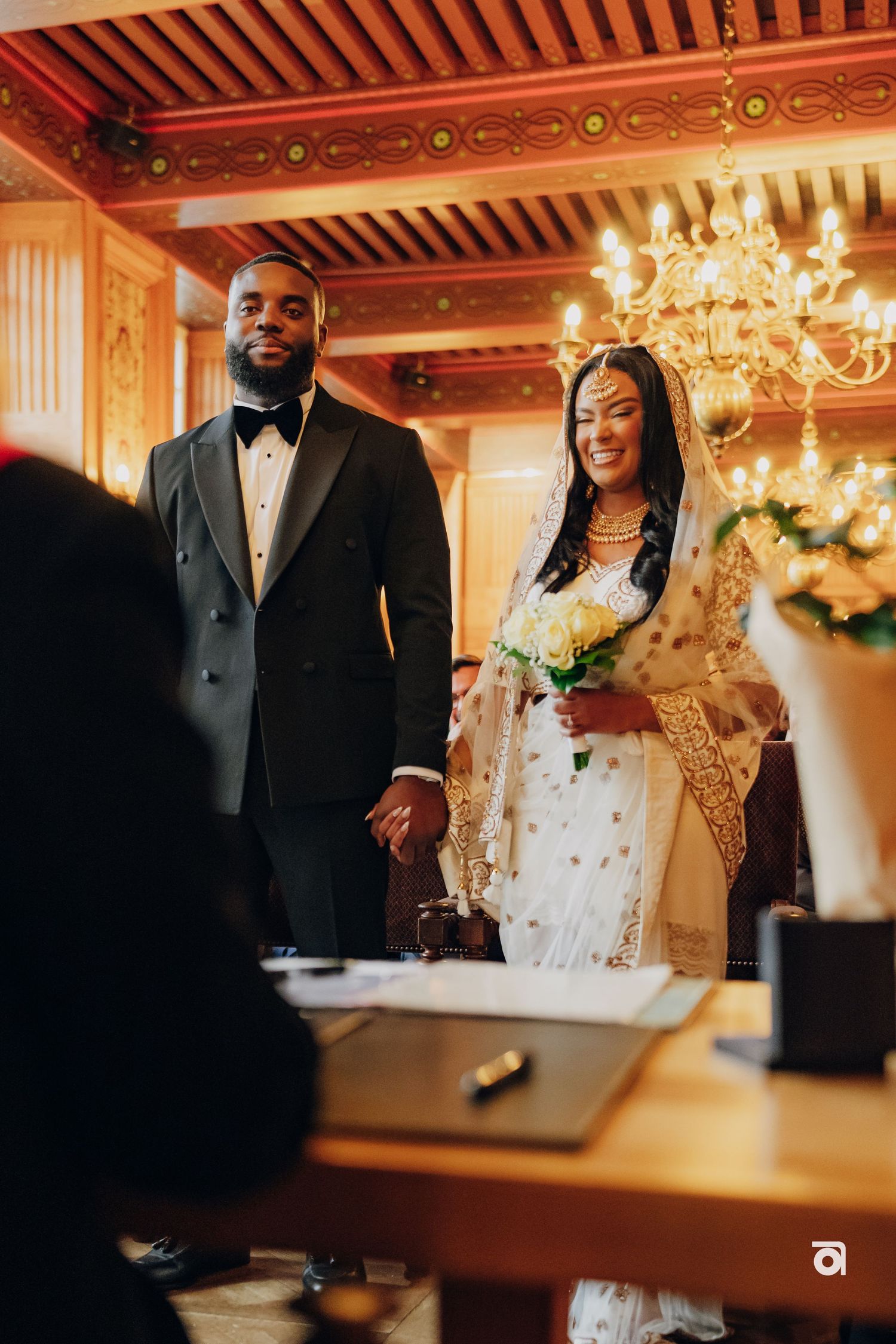 A wedding ceremony in an ornate room with a chandelier and decorative wooden ceiling.