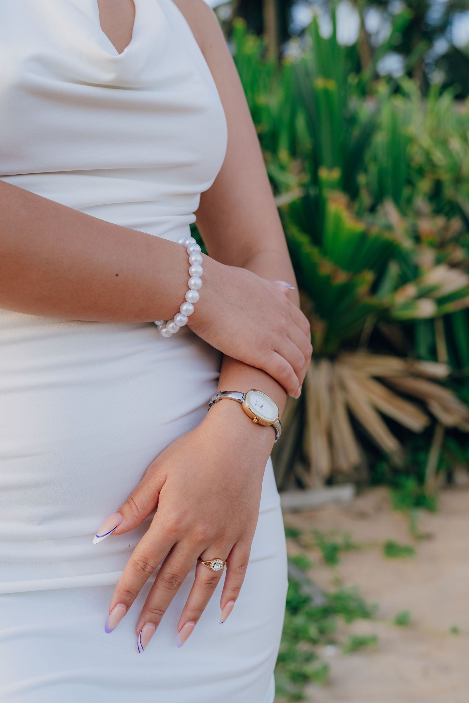 Bride in white dress showcasing pearl bracelet, gold watch, and diamond engagement ring against tropical greenery.