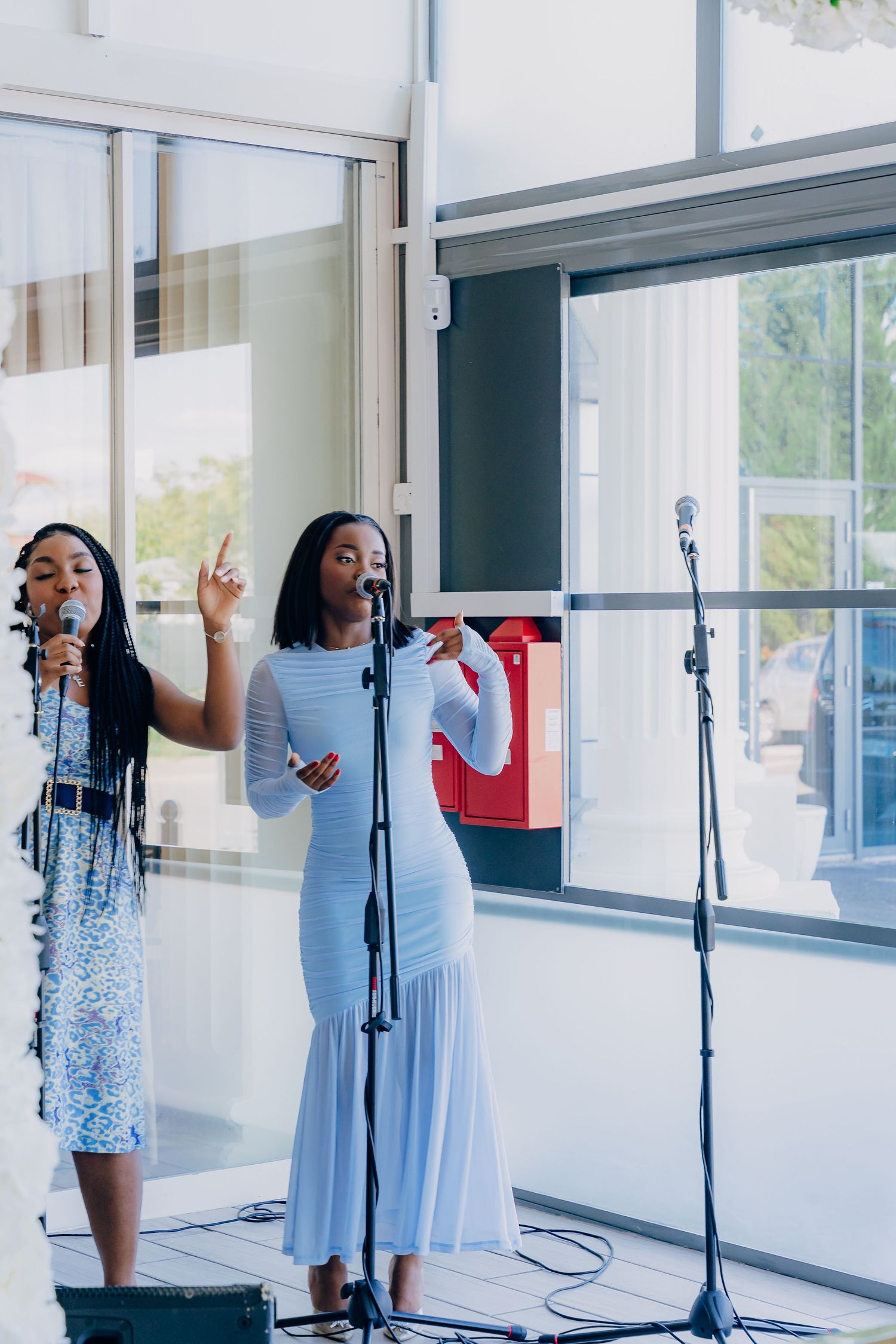 Wedding singers perform at microphone stands in a bright indoor venue with large windows.