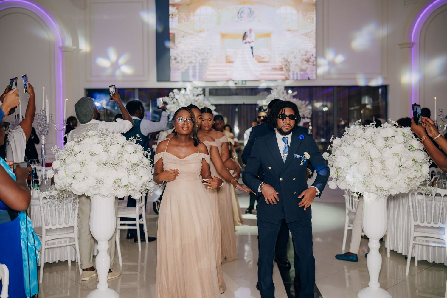 Wedding celebration in an elegant white reception hall with tall floral centerpieces and guests dancing.