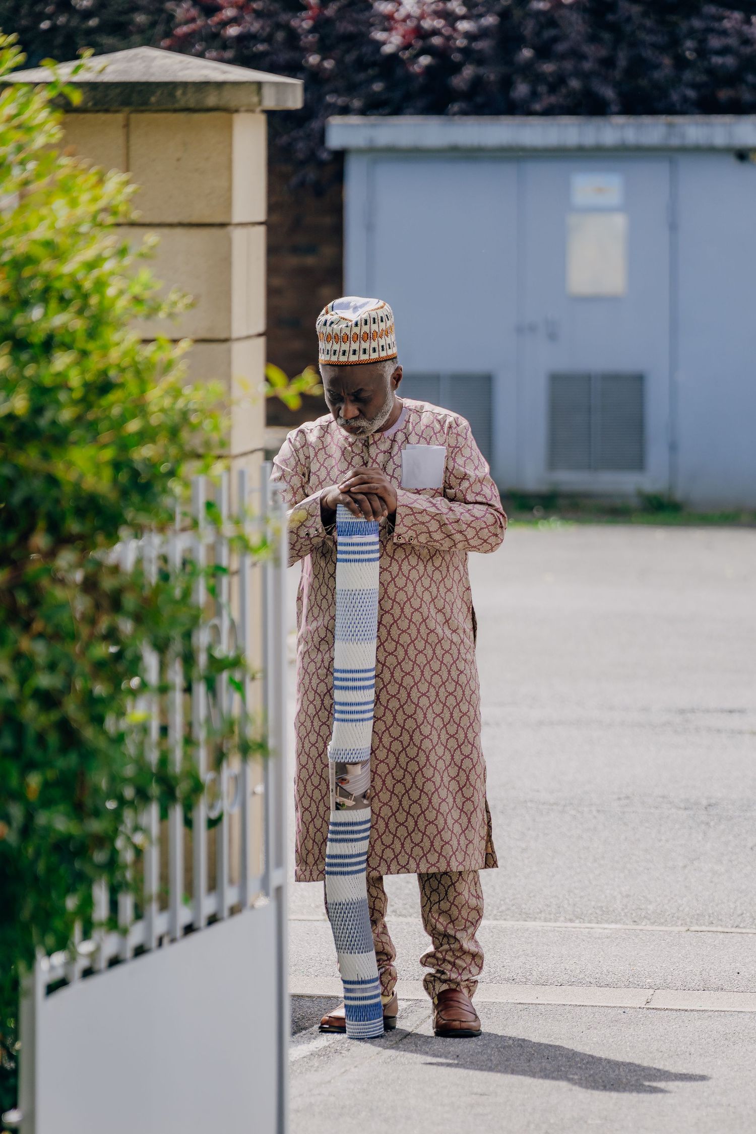 A person wearing traditional brown patterned clothing and prayer cap stands outdoors near a white picket fence.