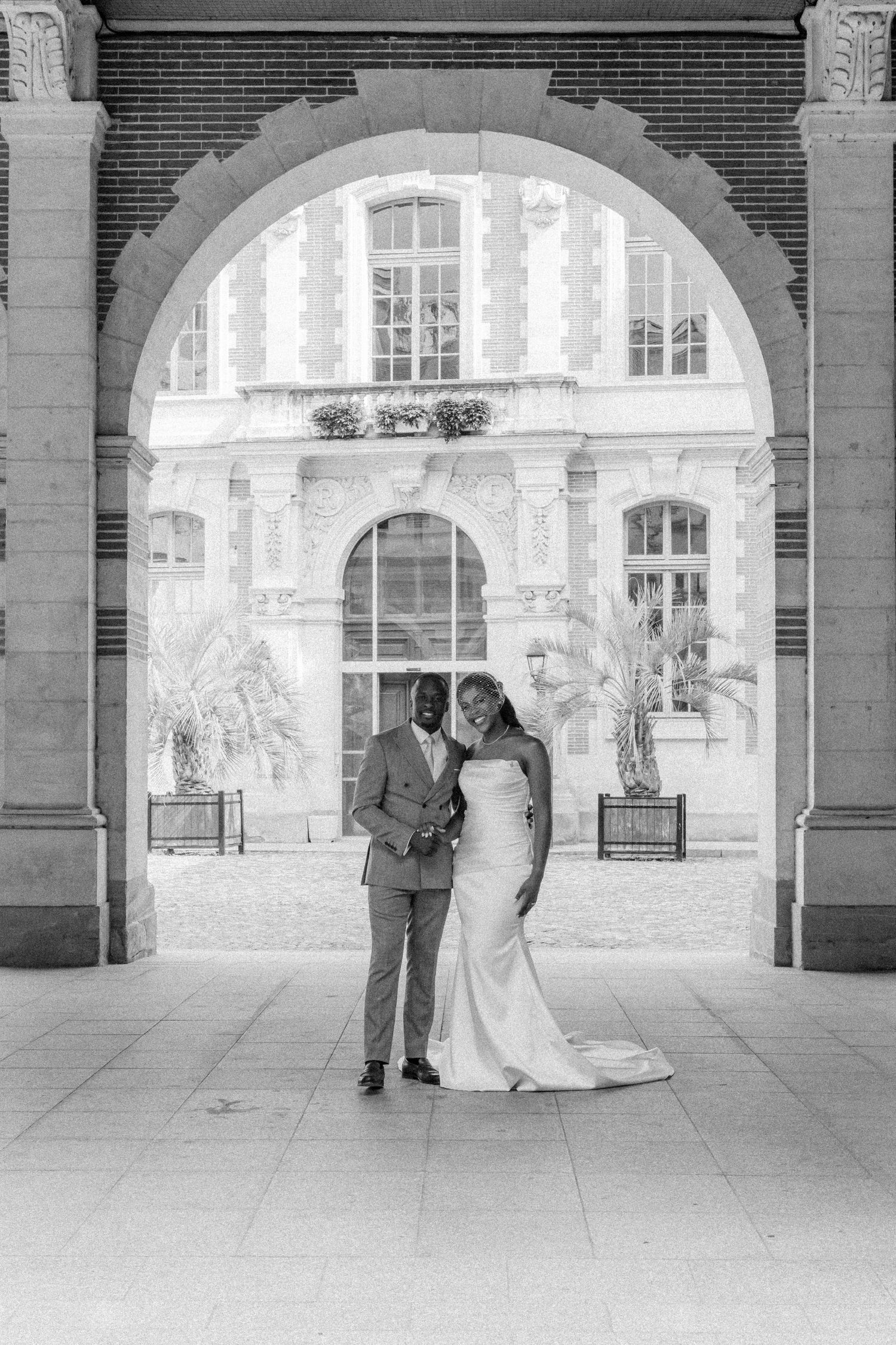 A couple stands together under an elegant archway in a historic building courtyard, captured in black and white.