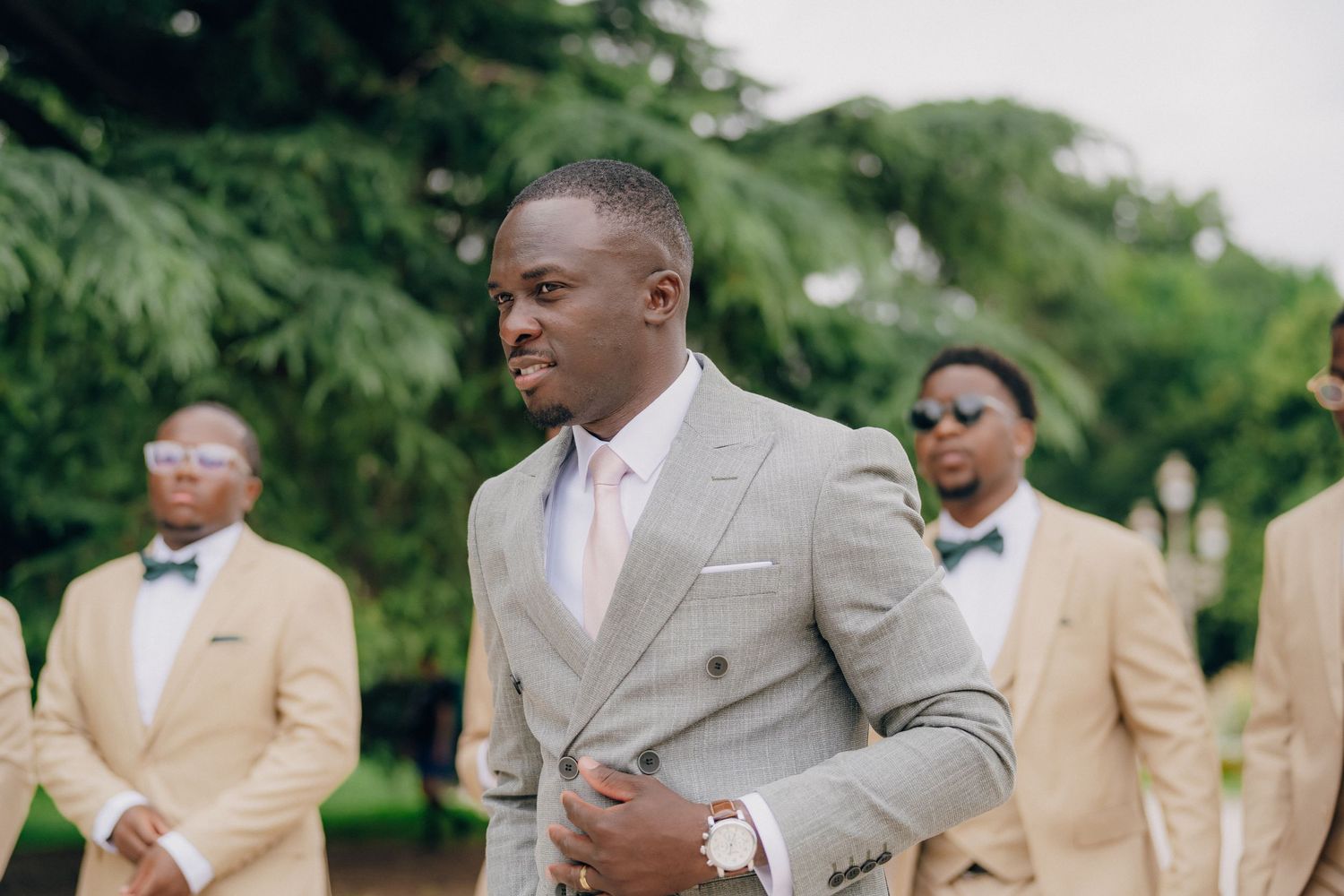 Wedding party members in tan and gray suits gather outdoors for formal photos during a summer celebration.
