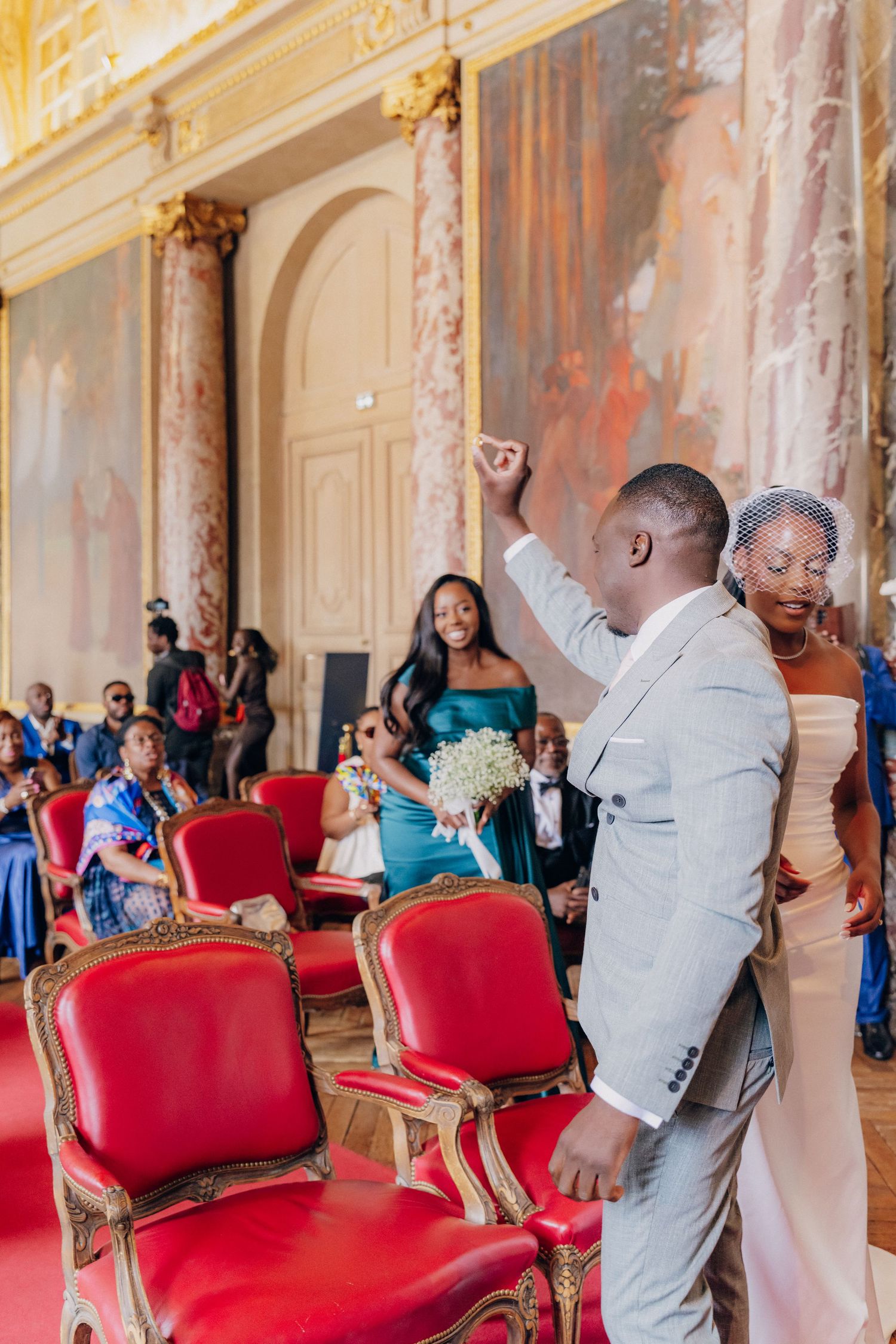 Wedding celebration in an ornate palace hall with red velvet chairs and decorative wall murals.
