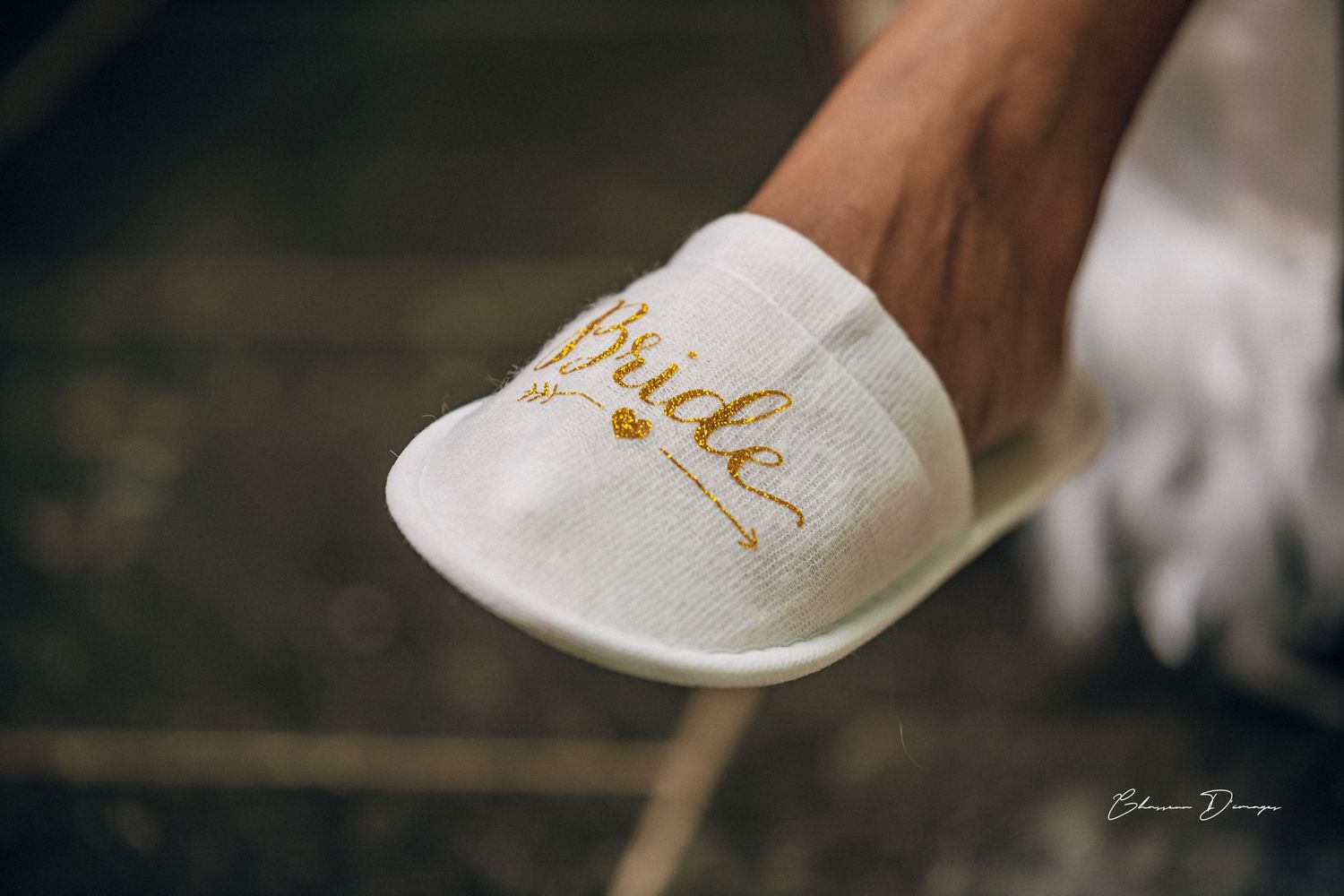 White bridal slipper with gold 'Bride' text detail against dark floor.