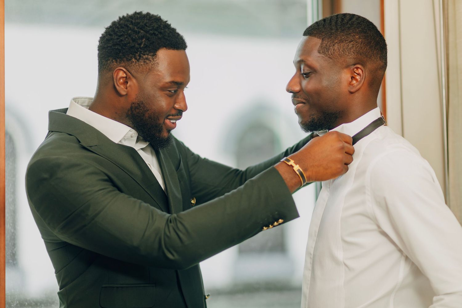 Two men in formal attire share a candid moment adjusting a bow tie before a special event.