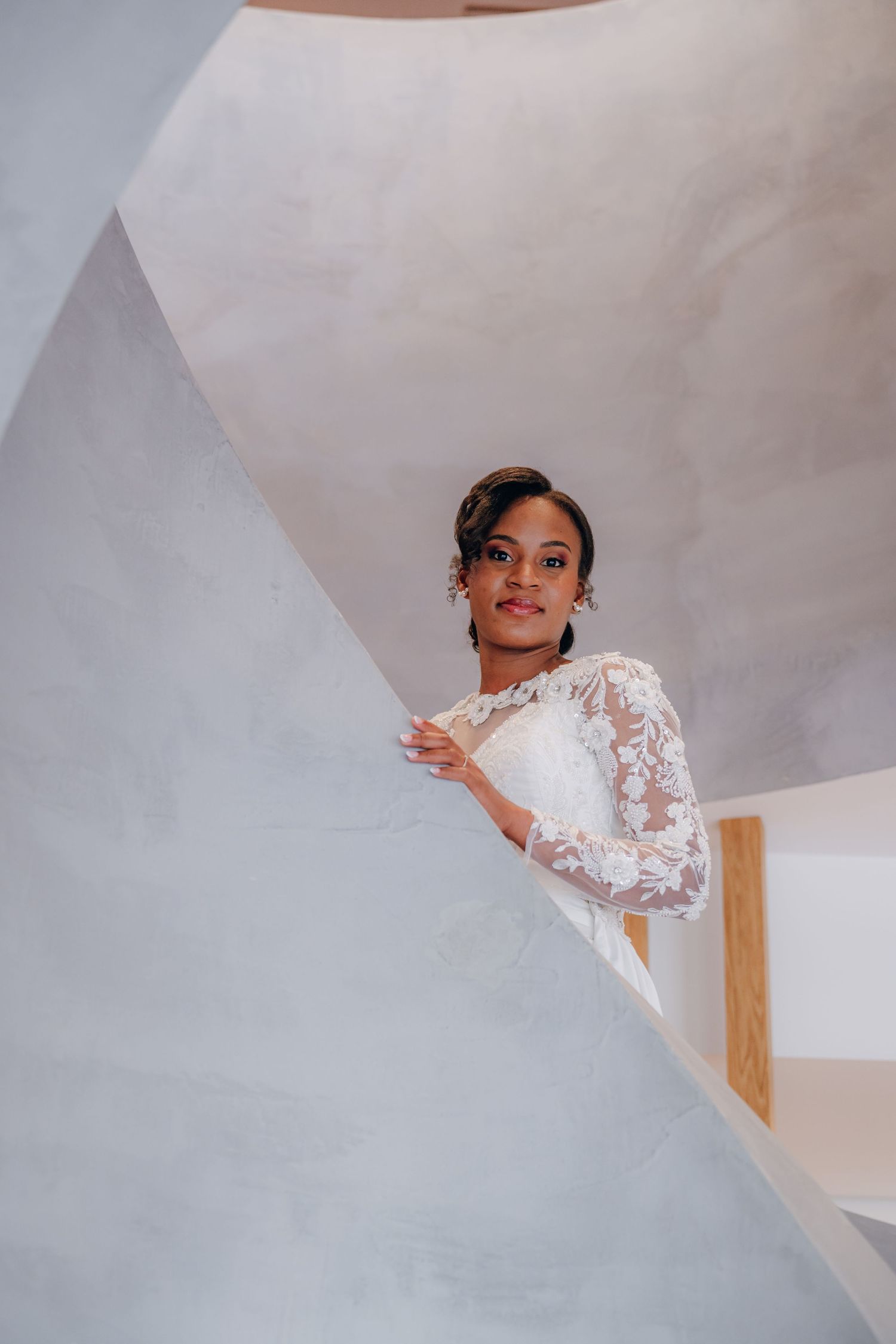 Someone in an elegant white lace wedding dress with long sleeves poses on a modern staircase in soft lighting.