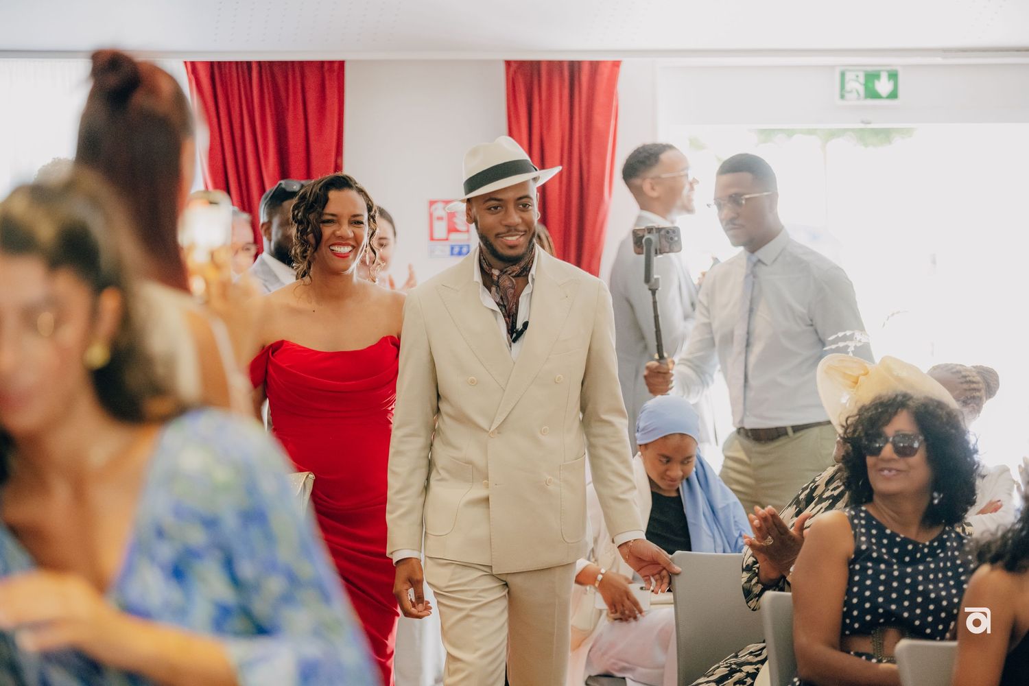 Wedding party attendees in formal attire walk down the aisle, with someone wearing a beige suit and fedora in focus.