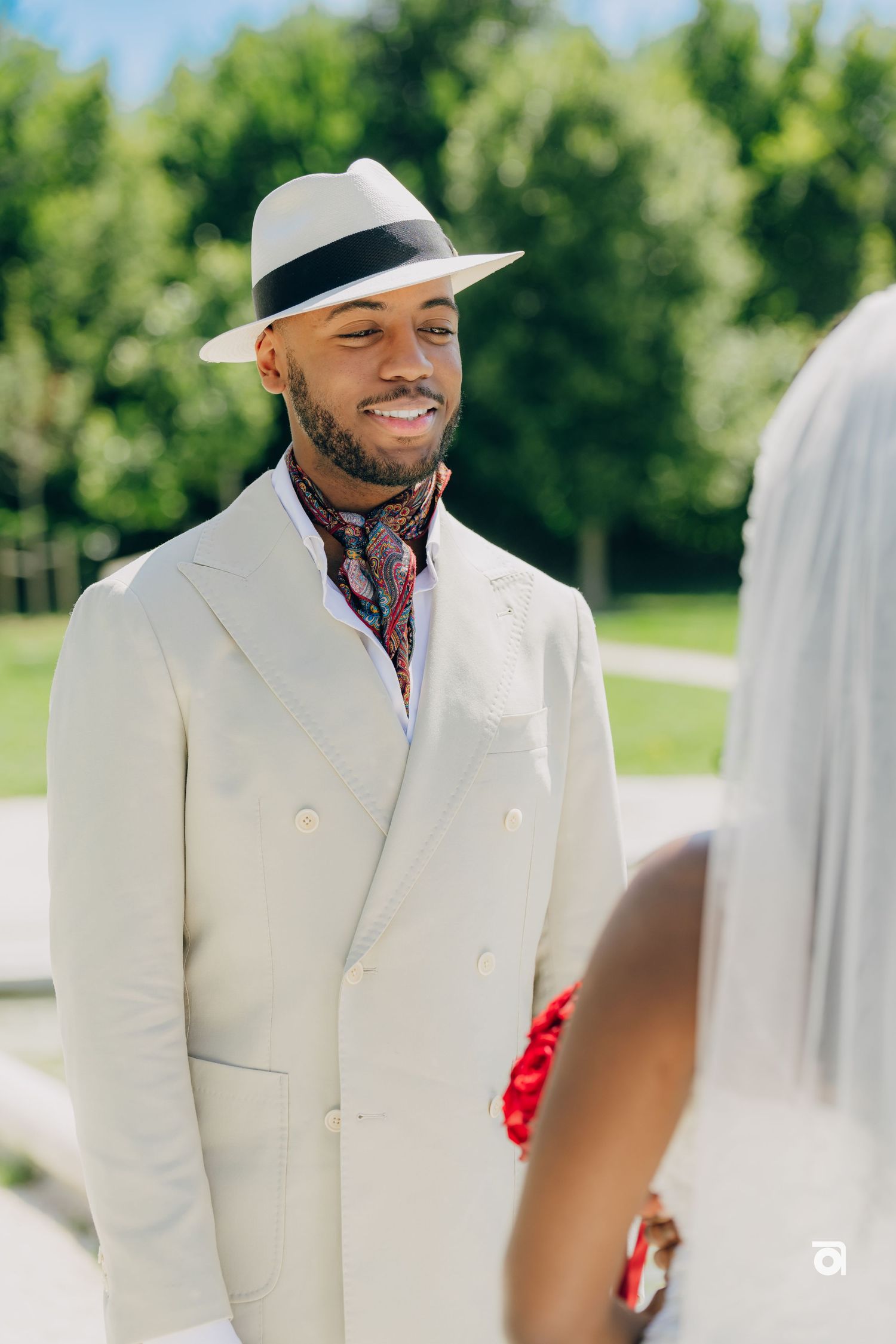 A groom in a white suit and fedora hat smiles during an outdoor wedding ceremony.