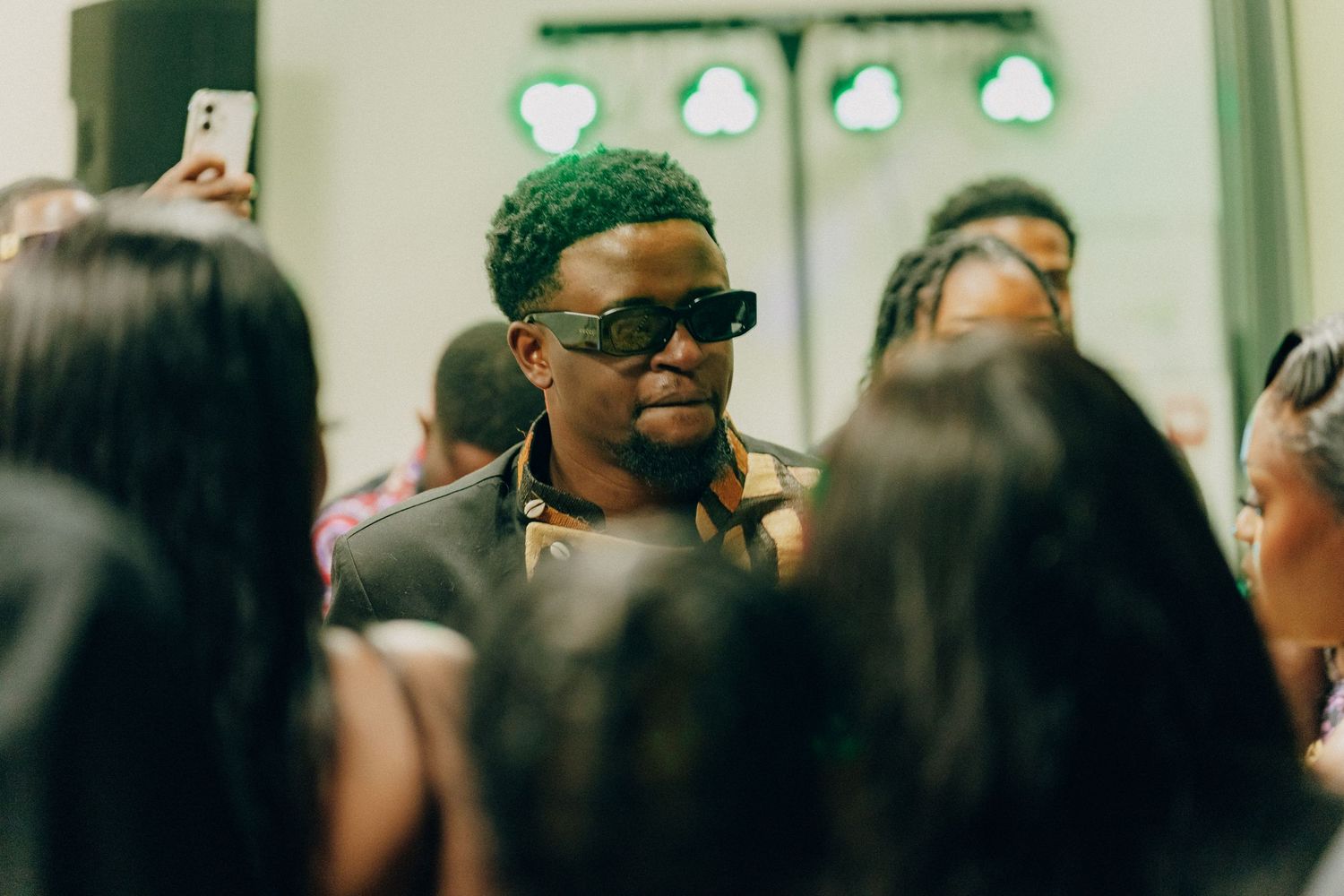 Someone wearing sunglasses and green hair smiles while surrounded by people at an indoor gathering.