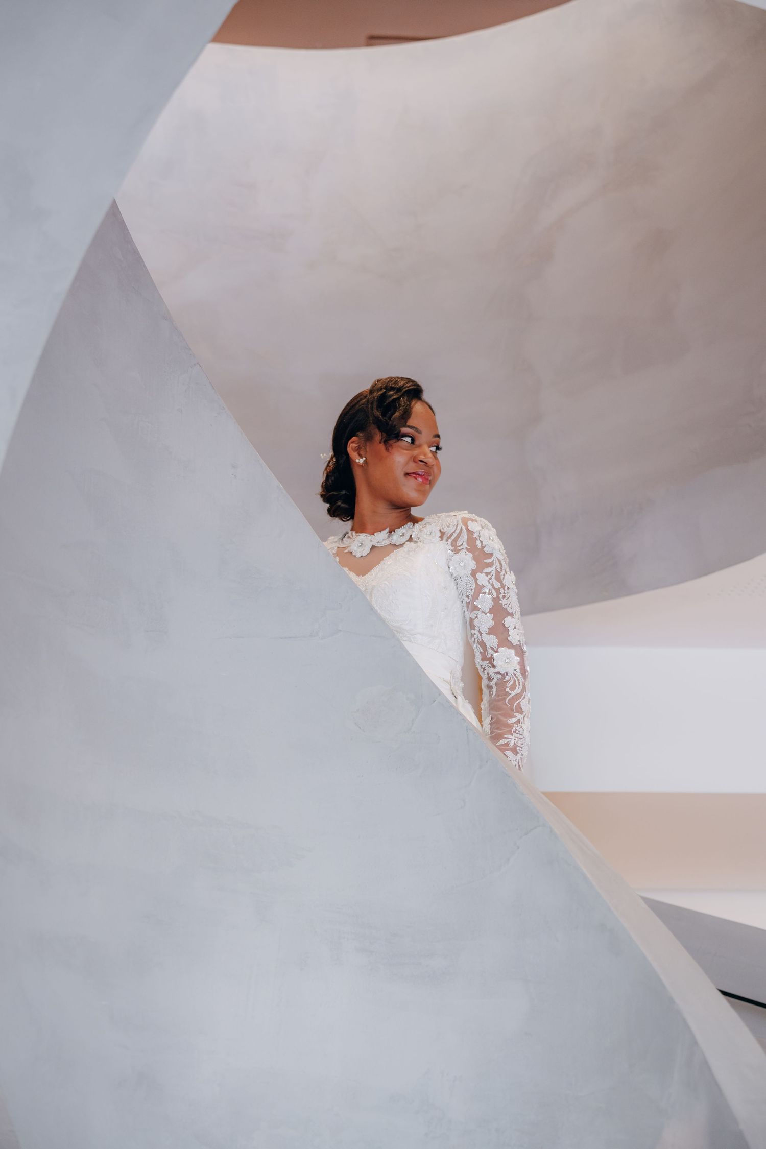 A bride in a white lace wedding dress stands elegantly on a modern white staircase with dramatic lighting.