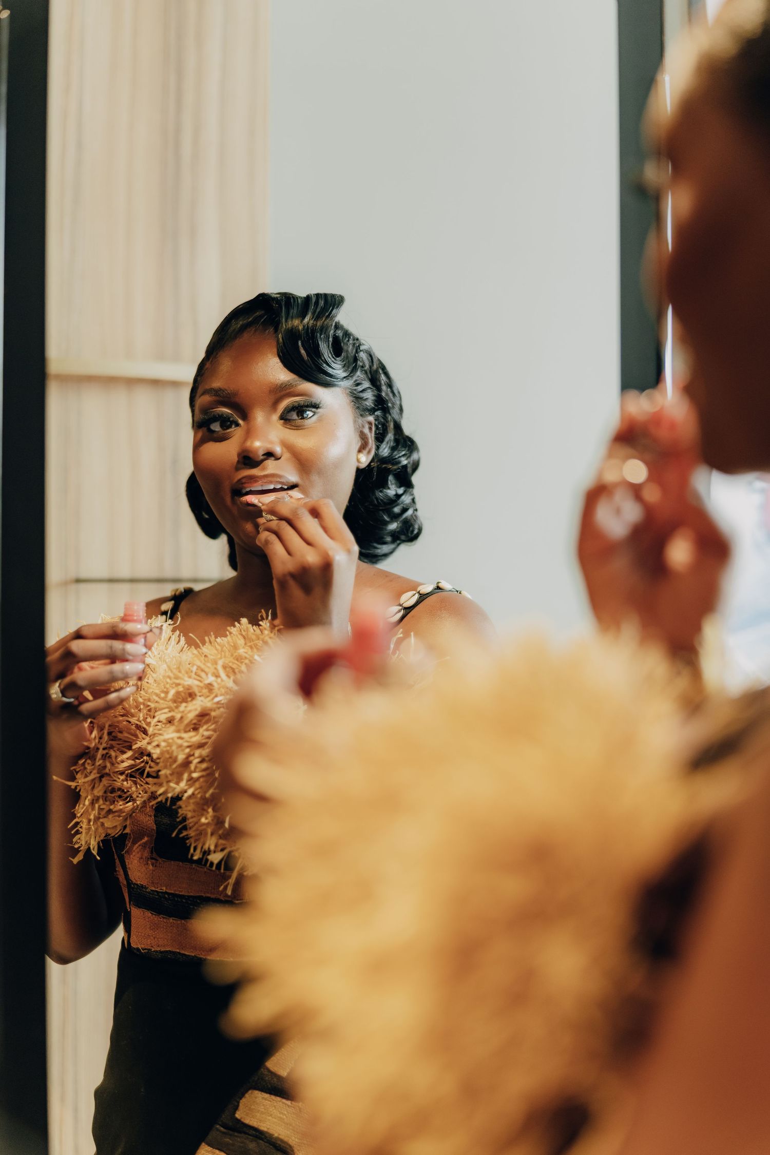 A person wearing elegant formalwear smiles while touching their face in a warm-lit room, reflecting in a mirror.