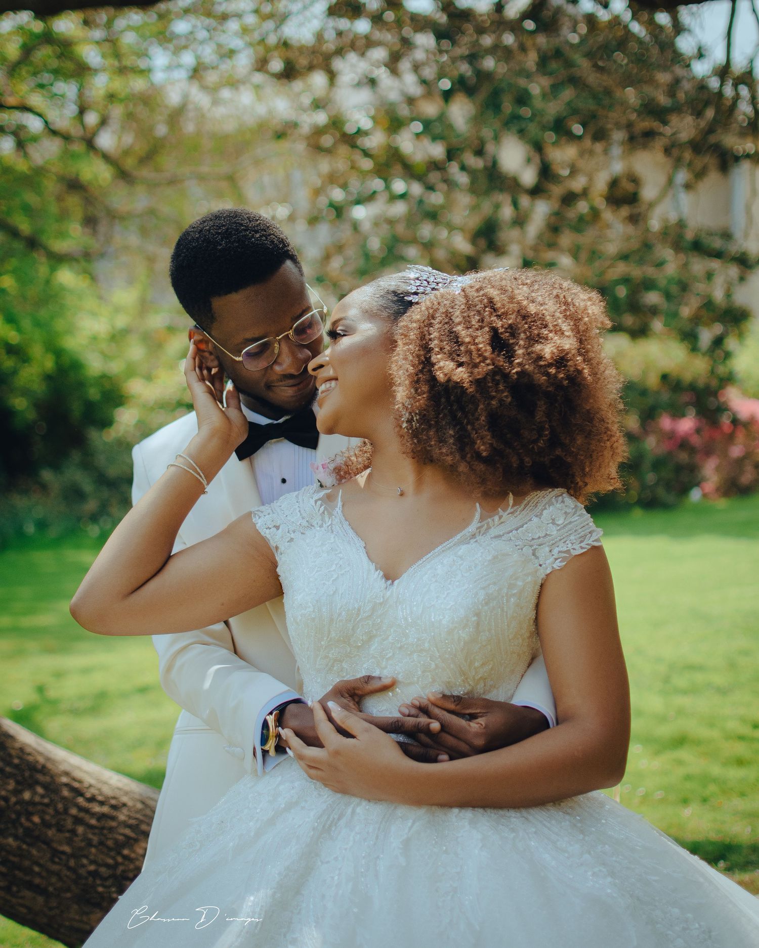 A loving couple in white wedding attire share an intimate moment in a sunlit garden setting.