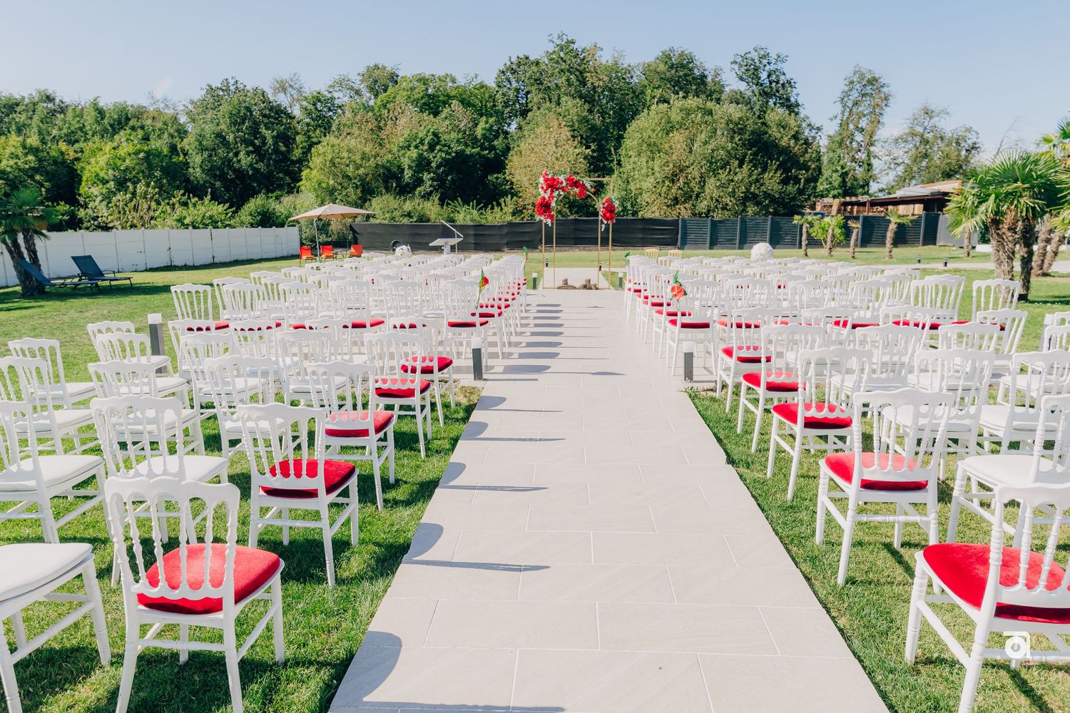 Outdoor wedding ceremony setup with white chairs and pink cushions leading to an altar on a sunny day.