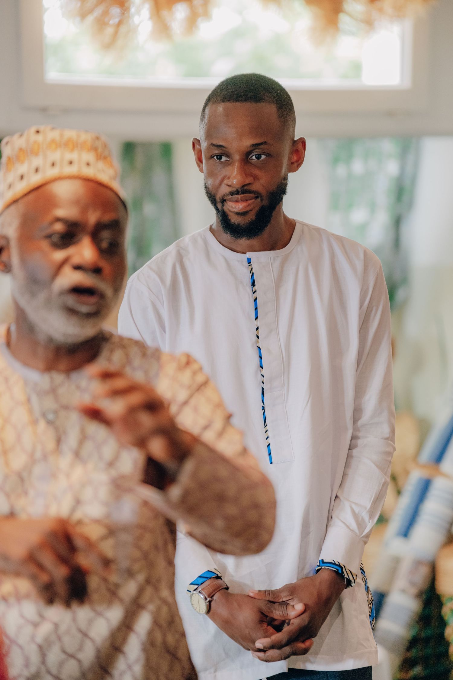 Two people in traditional and modern attire stand together in a bright indoor setting.