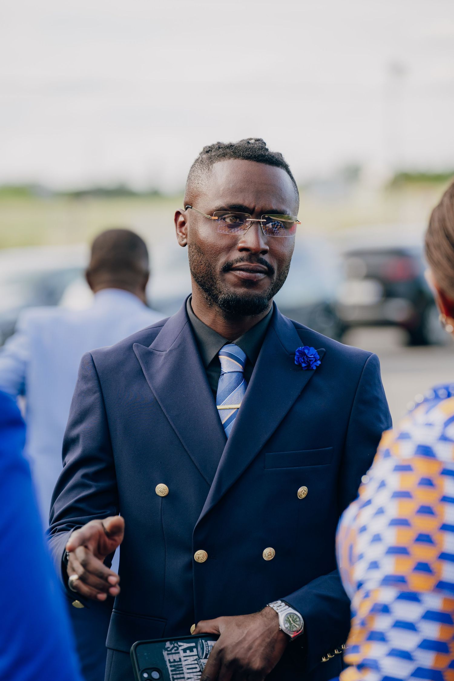 Man in navy blazer with gold buttons and blue boutonniere smiling at outdoor event.
