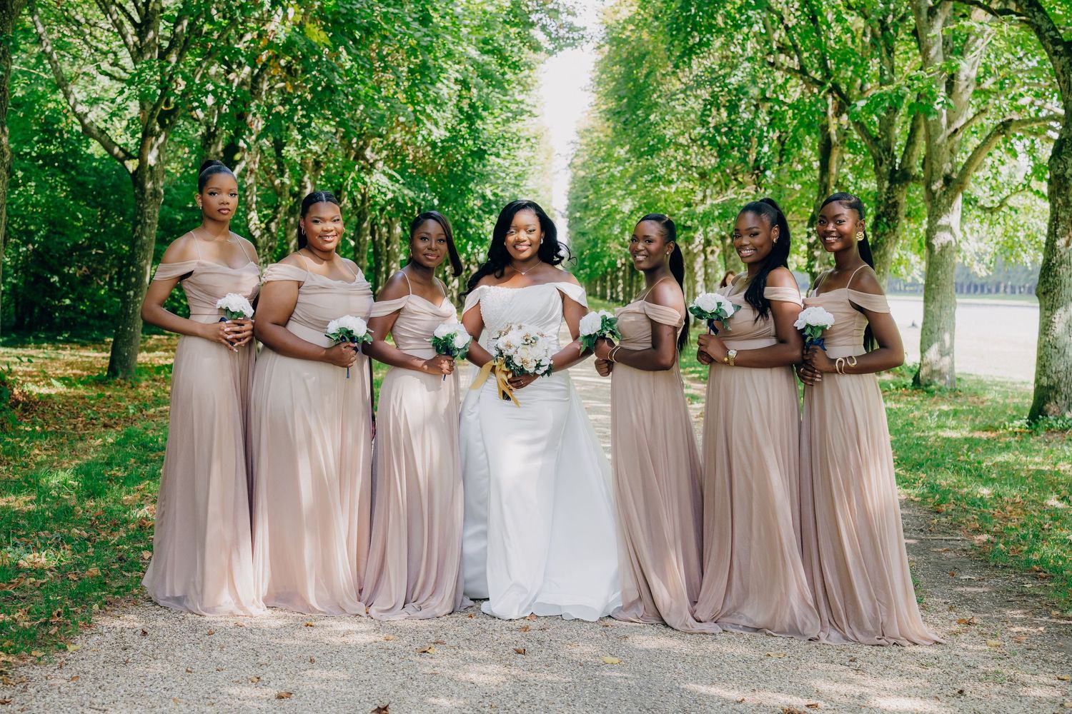 Bridesmaids in neutral dresses pose together on a tree-lined path during an outdoor wedding.