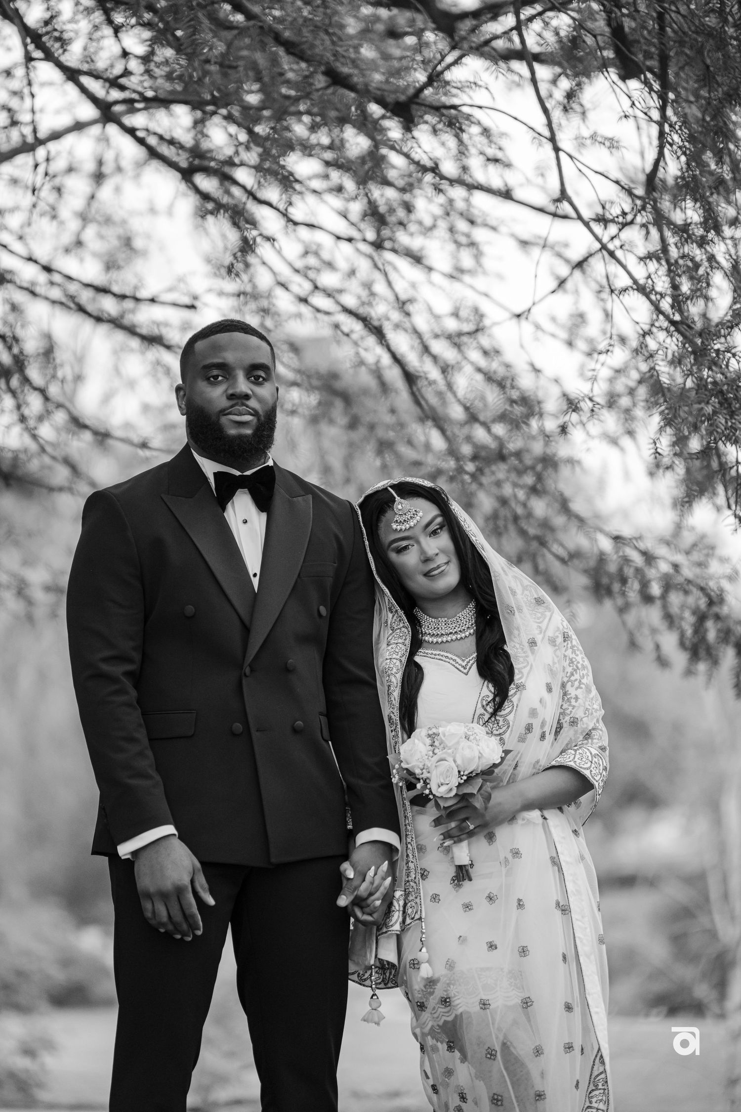 Black and white outdoor wedding photo of a couple in formal attire holding hands under tree branches.