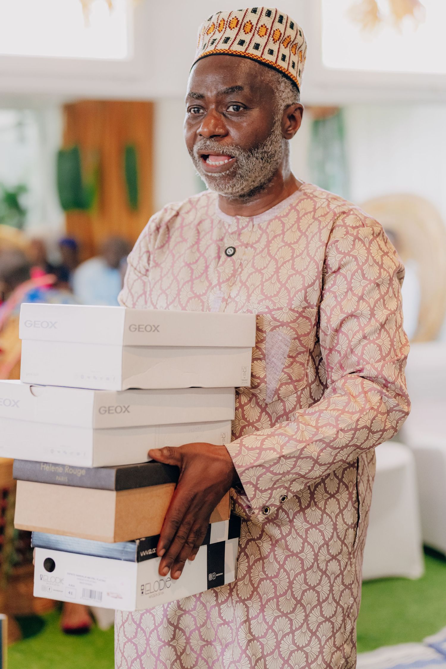 Person wearing traditional African attire holding stacked white gift boxes at an event.