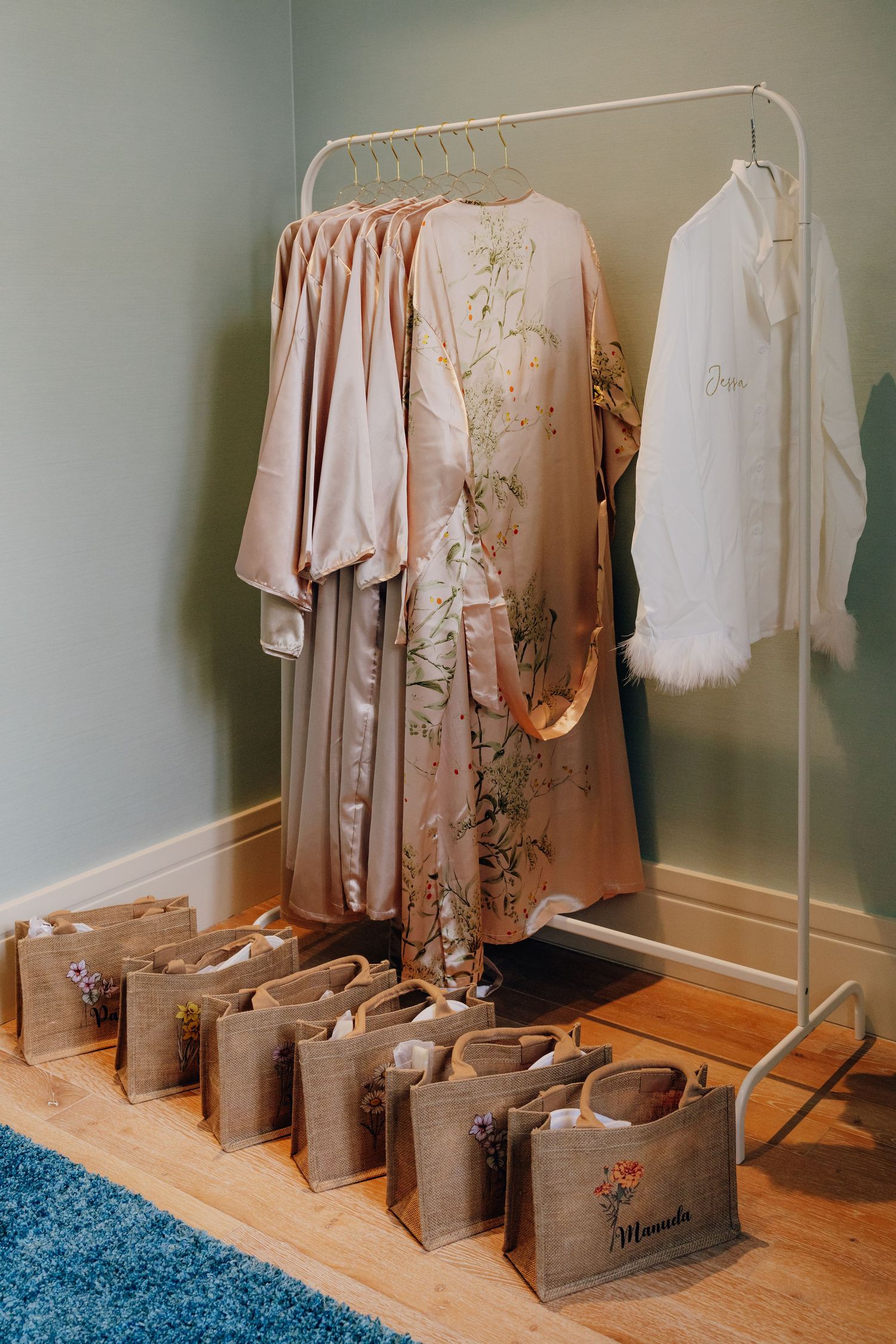 Wedding party favors and blush pink dresses displayed on a clothing rack with gift bags lined up below.