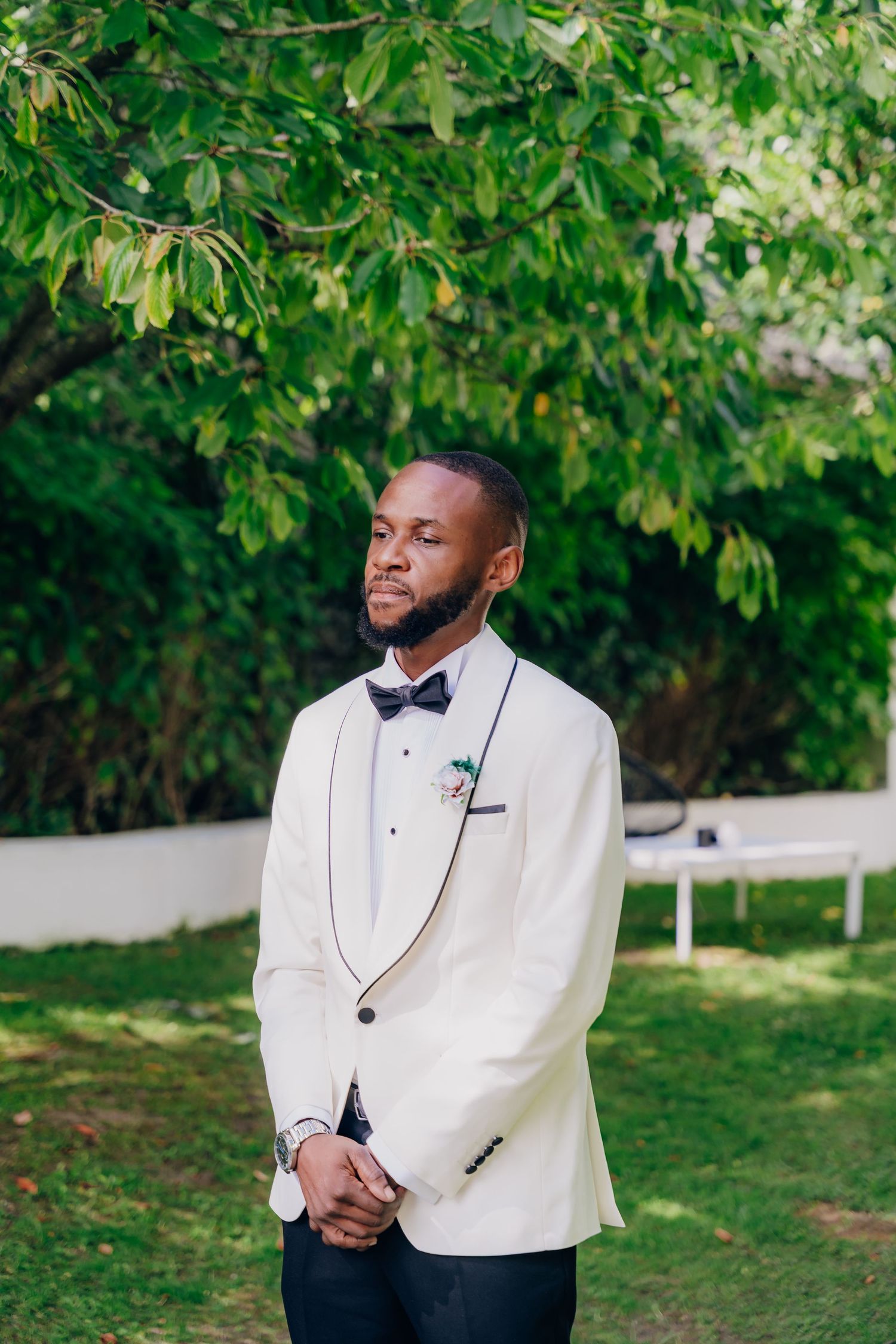 Groom in a white tuxedo jacket and black bow tie poses outdoors under green leafy trees.